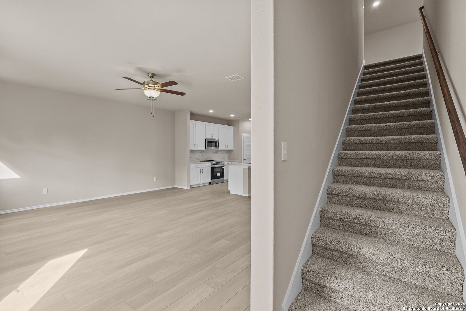 Open living room with hardwood floors, ceiling fan, modern white kitchen, and carpeted staircase in 2-story Davidson Homes The Douglas G, San Antonio