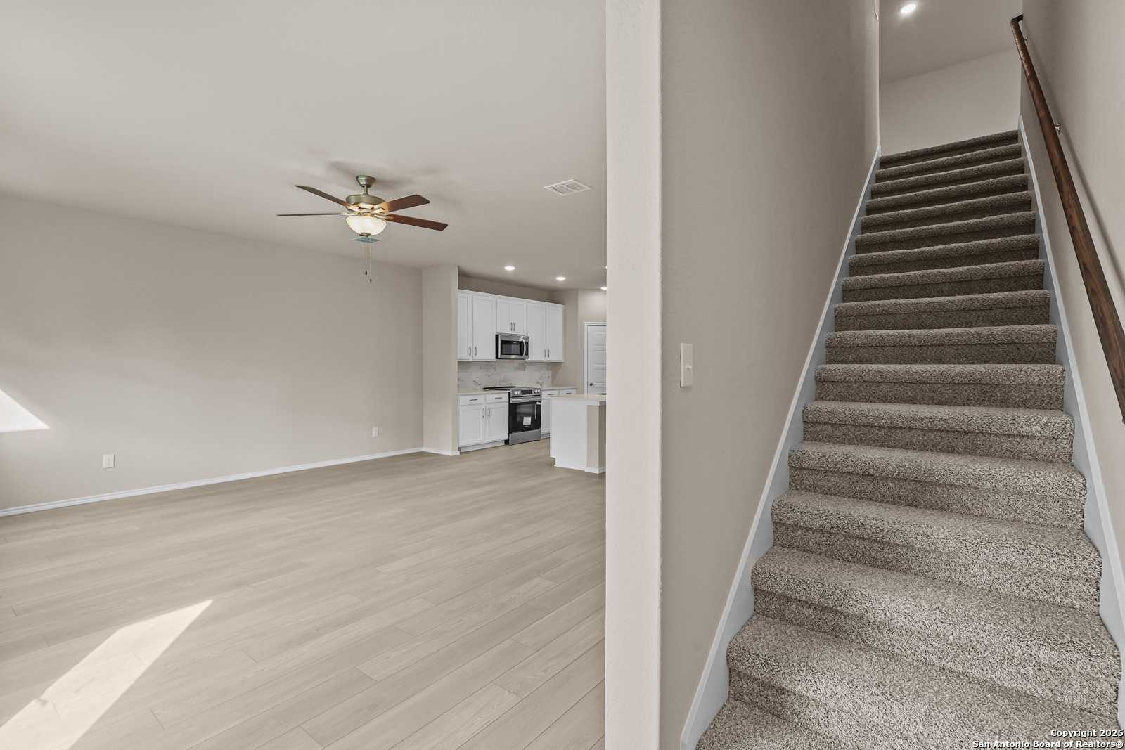 Open living room with hardwood floors, ceiling fan, modern white kitchen, and carpeted staircase in 2-story Davidson Homes The Douglas G, San Antonio