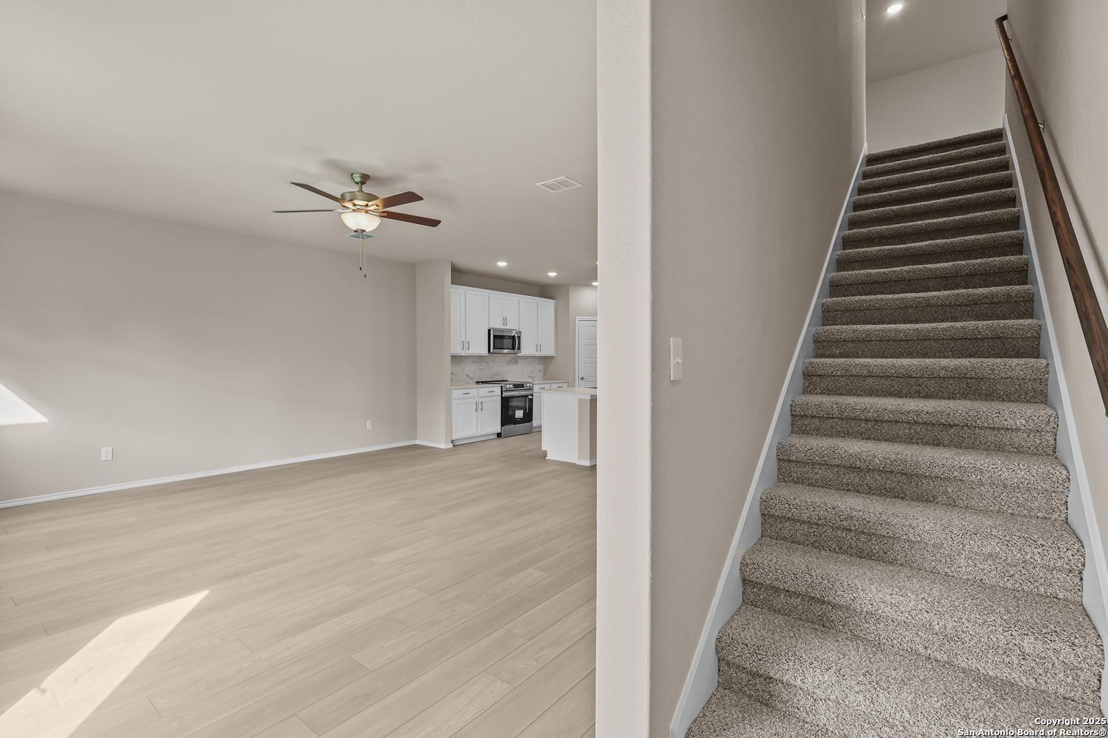 Open living room with hardwood floors, ceiling fan, modern white kitchen, and carpeted staircase in 2-story Davidson Homes The Douglas G, San Antonio