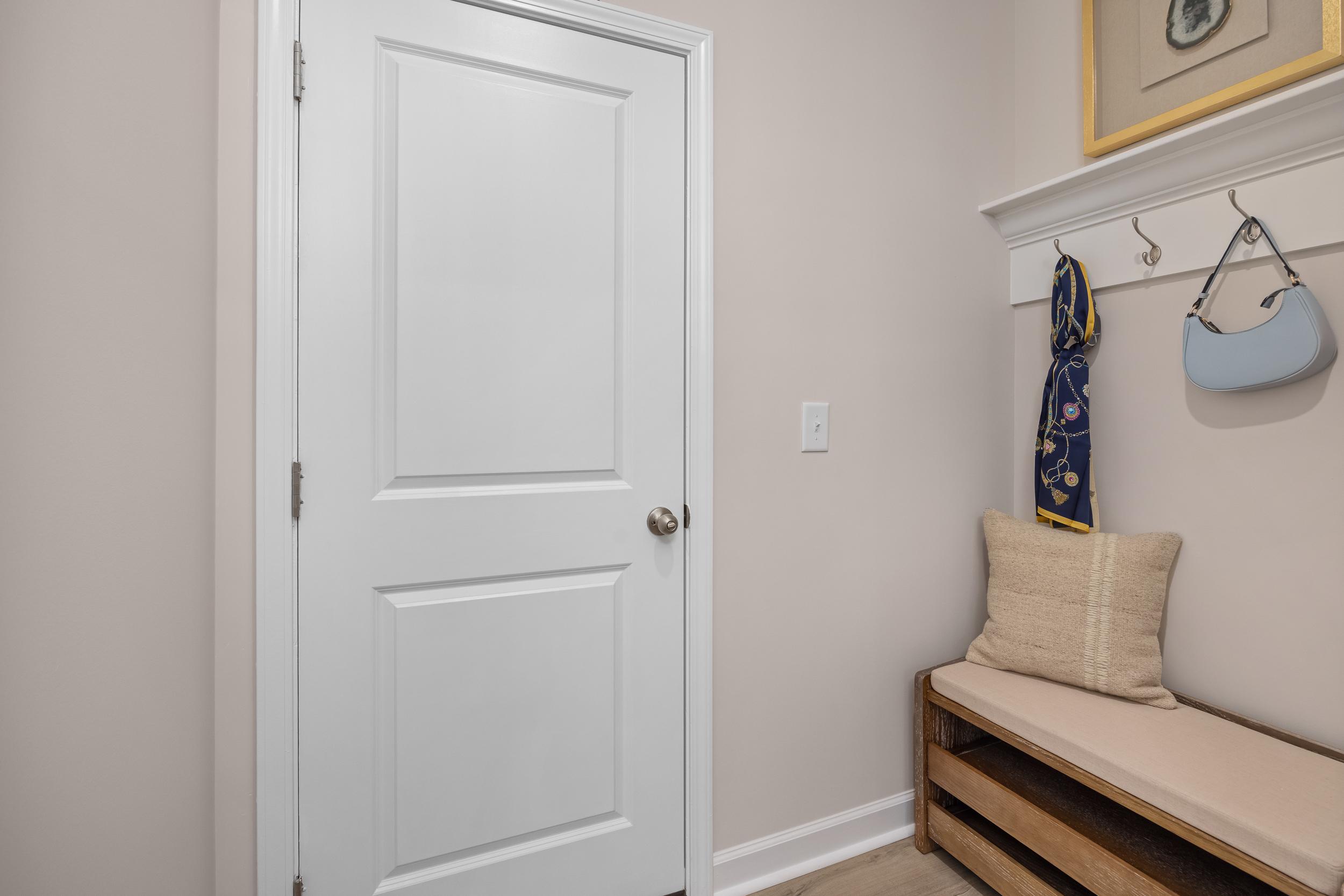 Cozy mudroom entry in The Hickory II with white door, built-in bench, coat hooks, scarf, handbag, and beige walls by Davidson Homes