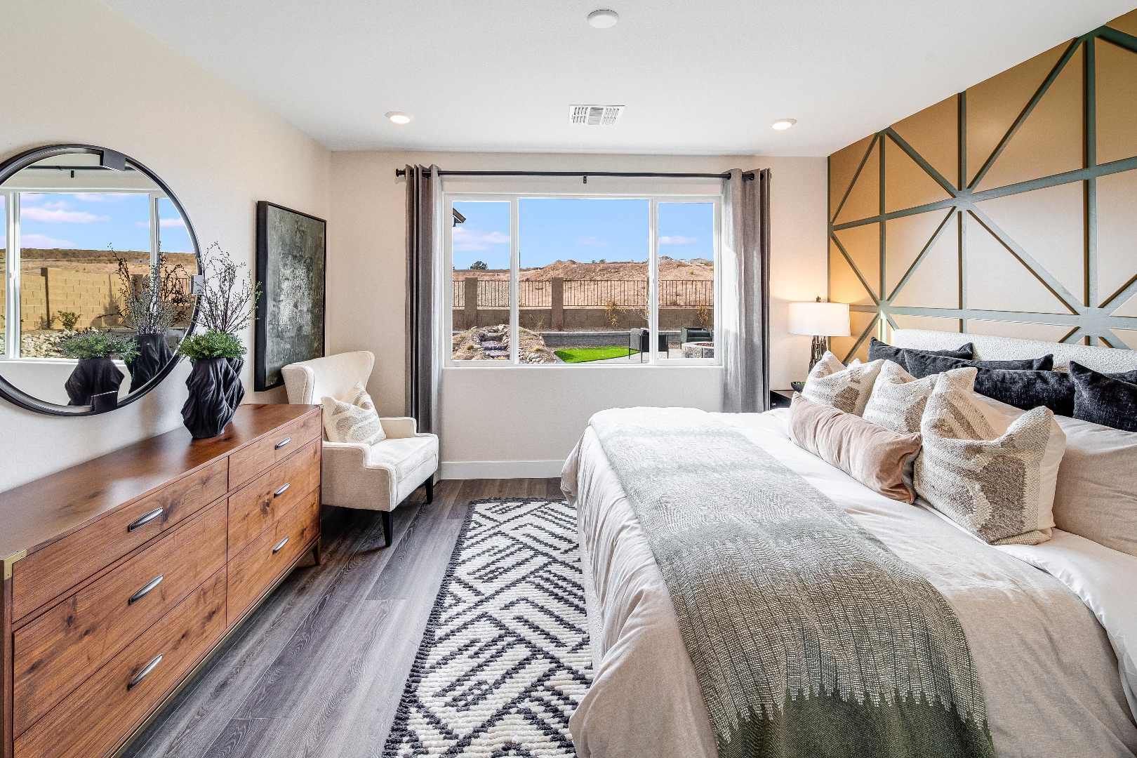 Spacious master bedroom in The Boulder II with king bed, wooden dresser, geometric gold wall, round mirror, and desert view window