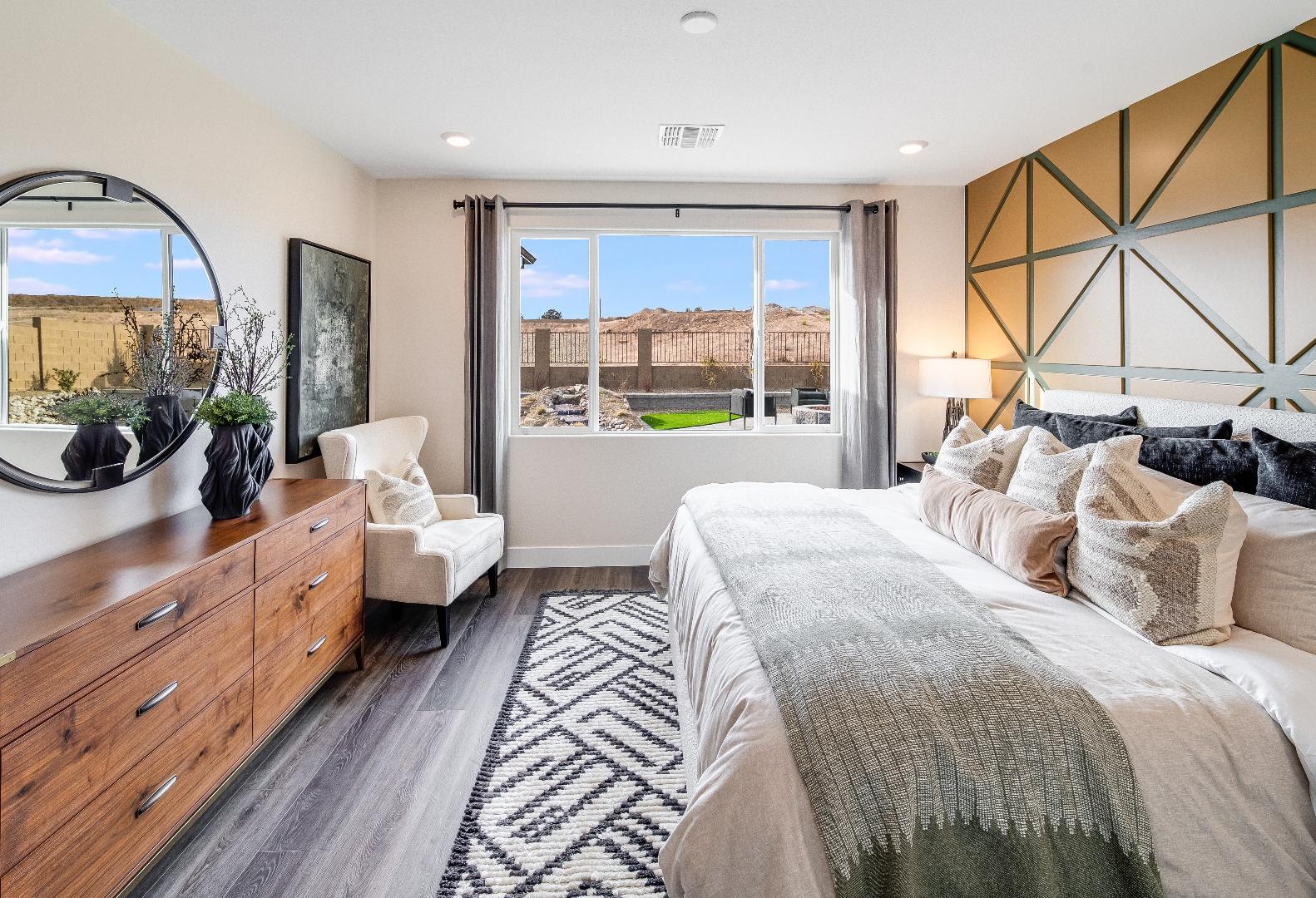 Spacious master suite in The Boulder II with king bed, geometric gold wall, wooden dresser, accent chair, and desert-view window