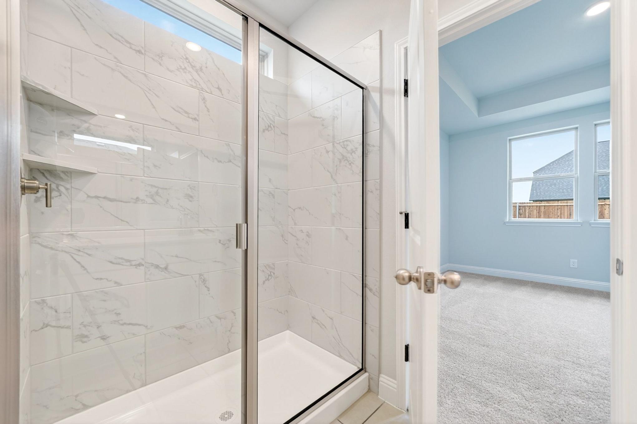 Walk-in shower with marble-look tile in The Redbud master suite featuring a view into the spacious primary bedroom.