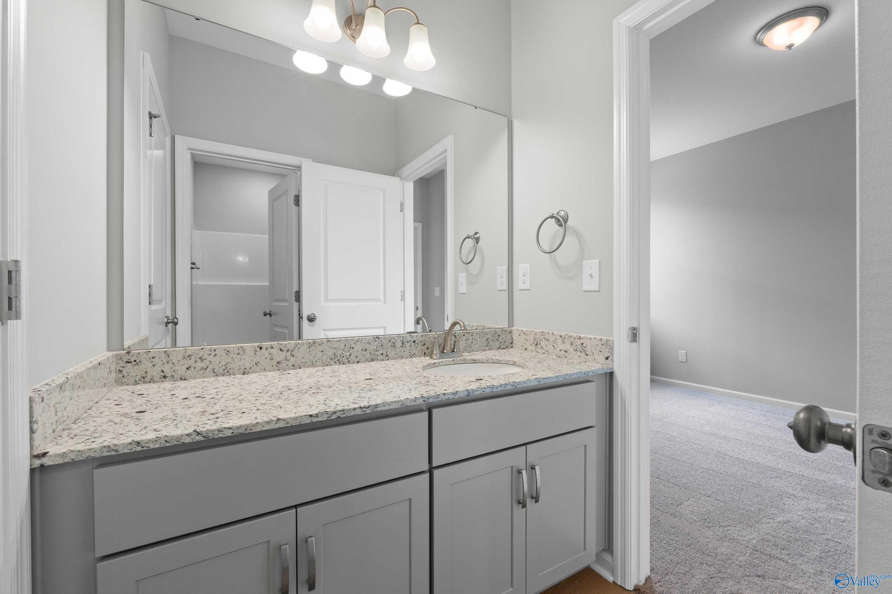 Modern master bathroom with granite double vanity, gray cabinets, and adjacent carpeted bedroom in Davidson Homes The Orion, Athens AL