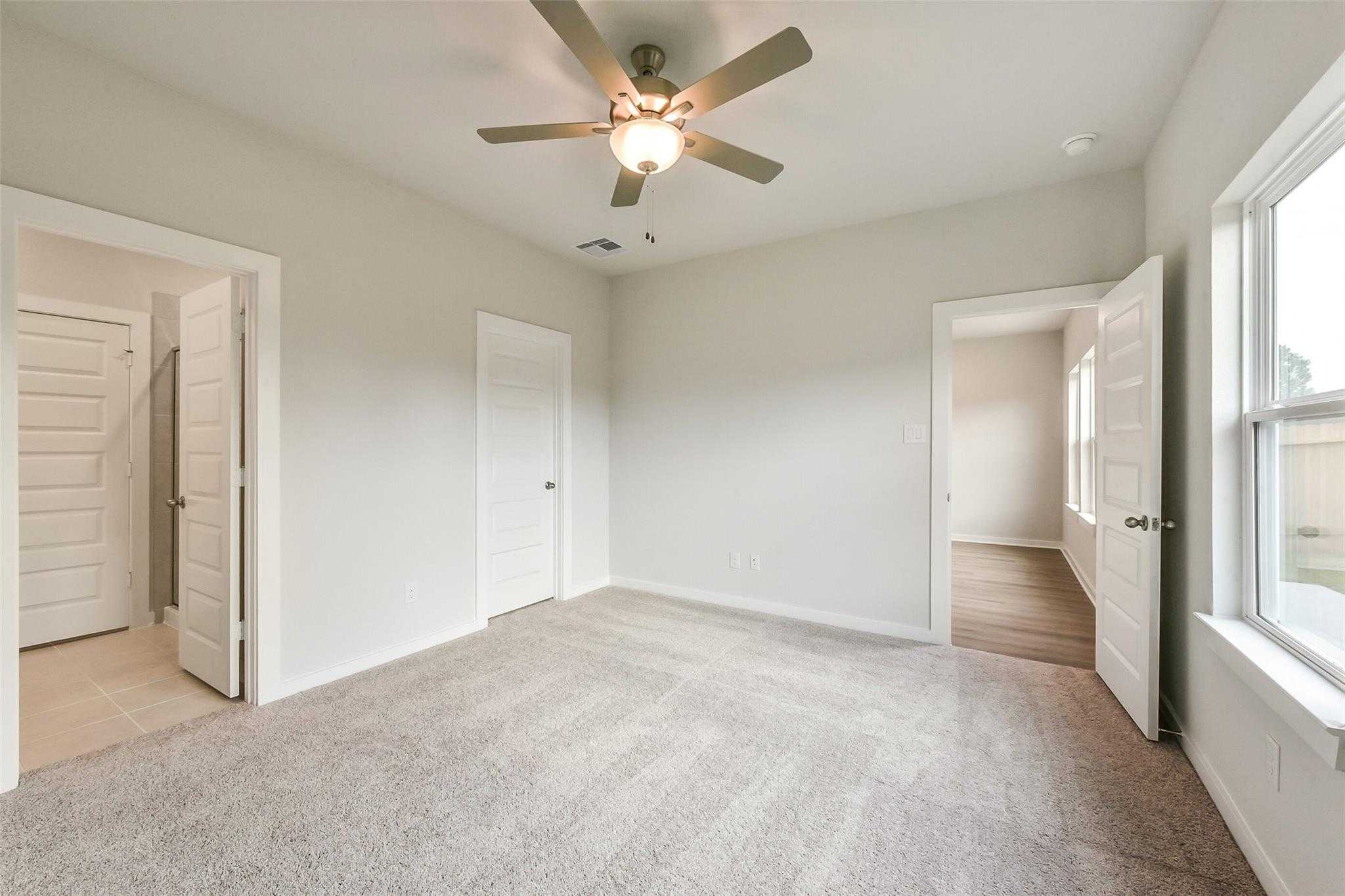 Bright bedroom with ceiling fan, beige walls, carpet flooring, and open bathroom door in Davidson Homes The Frio G, Dayton Texas
