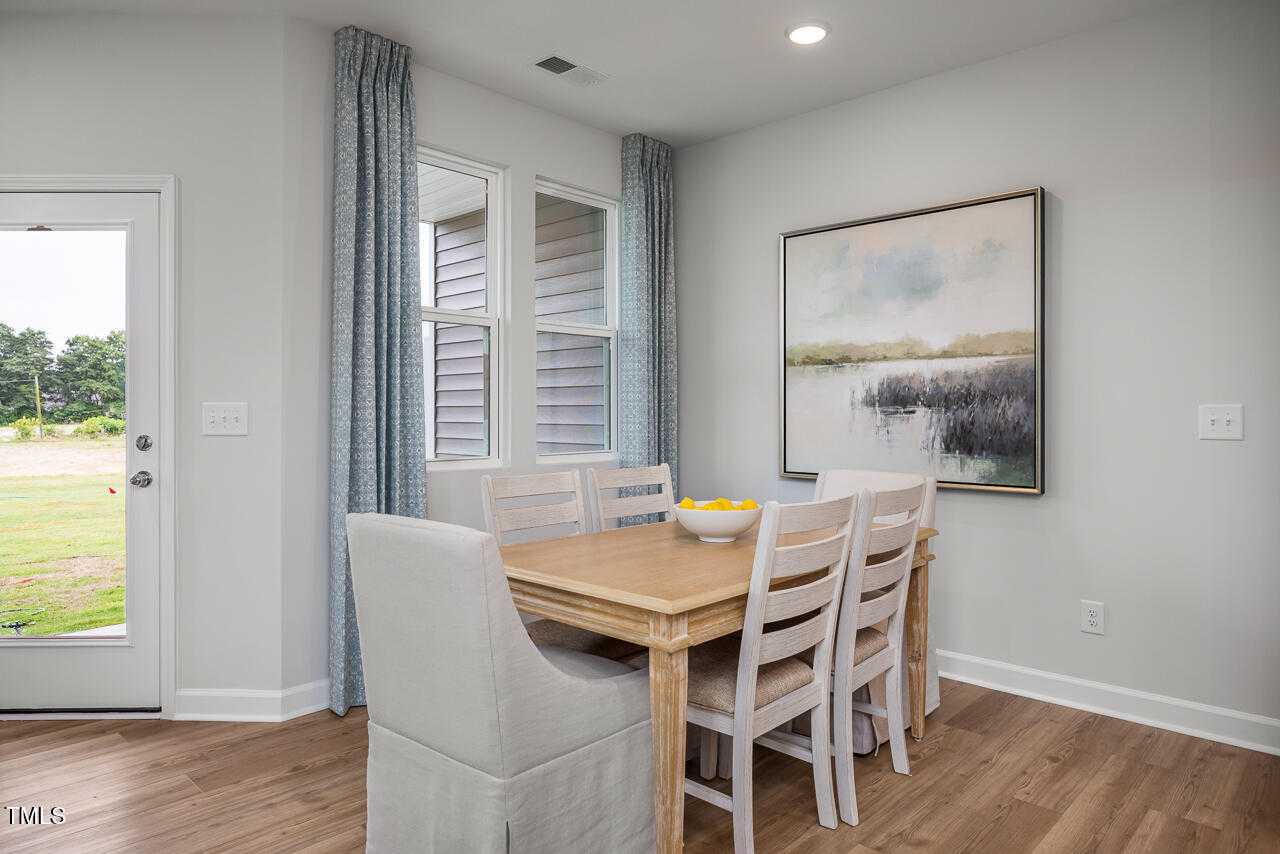 Bright dining room with wooden table, white upholstered chairs, fruit bowl, and landscape painting overlooking yard in Davidson Homes The Graham, Fuquay-Varina, NC