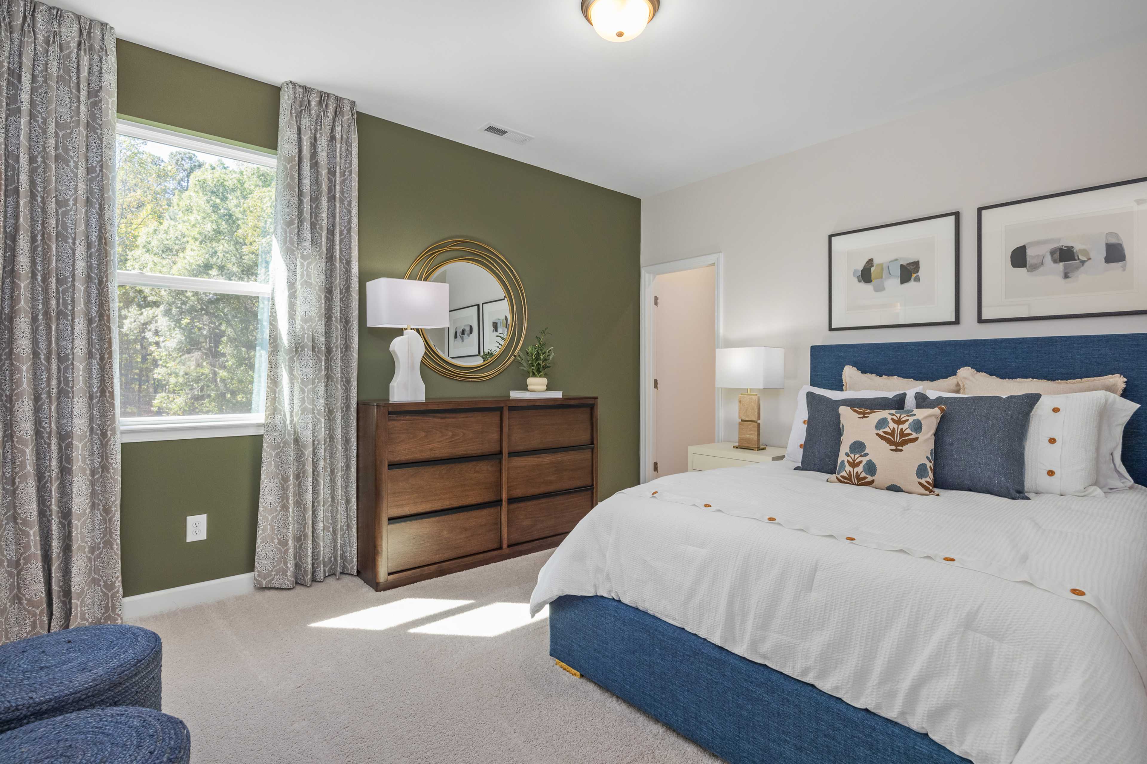 Spacious primary bedroom at Sage on North Main in Wake Forest NC with green accent wall, blue upholstered bed, and window views