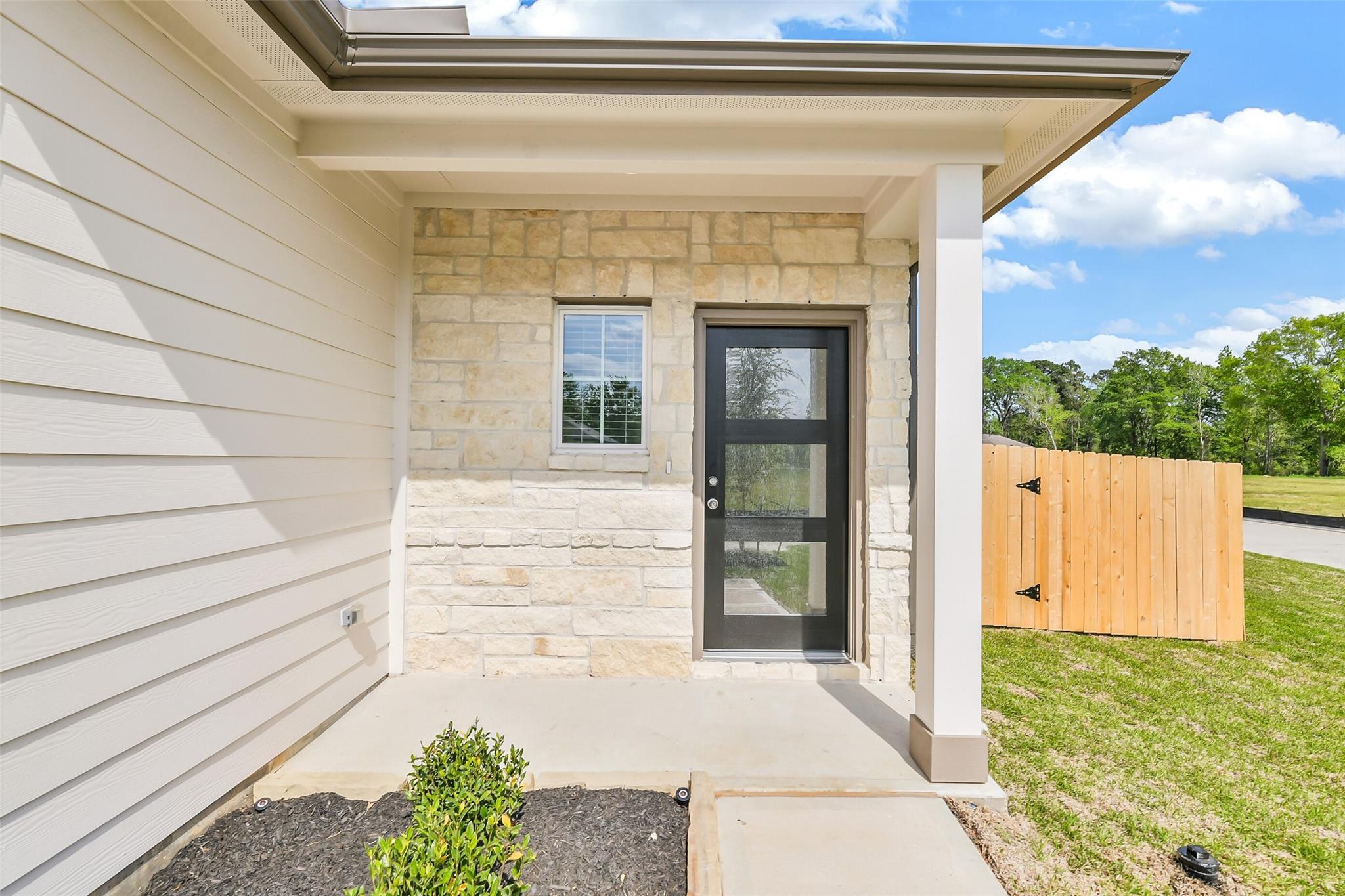 Modern front entrance with beige siding, stone accents, black glass door, and covered porch in Davidson Homes San Marcos E, Conroe Texas
