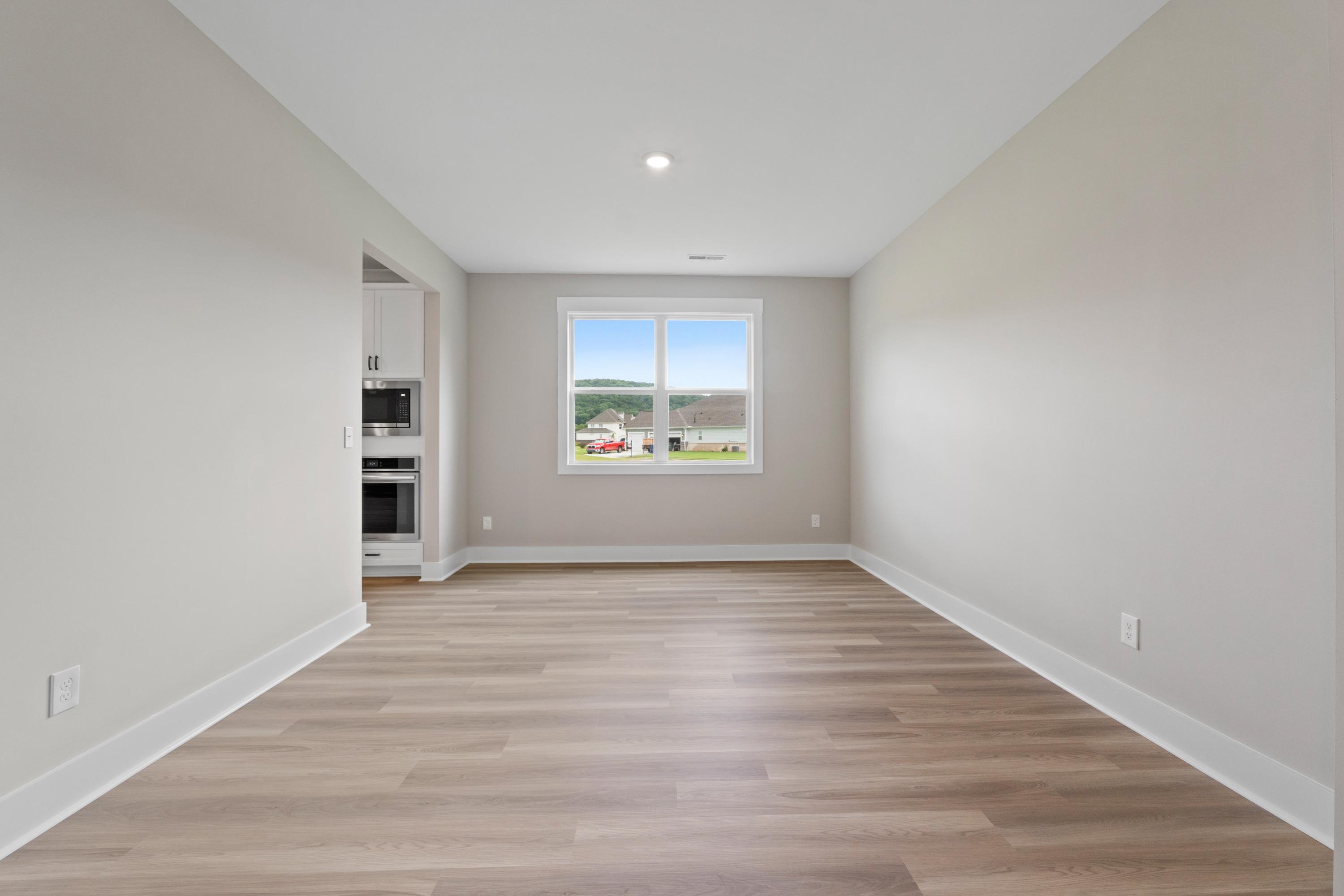 Spacious empty bedroom in The Oxford B by Evermore Homes with light hardwood floors, neutral walls, large window overlooking fields, adjacent kitchen cabinets