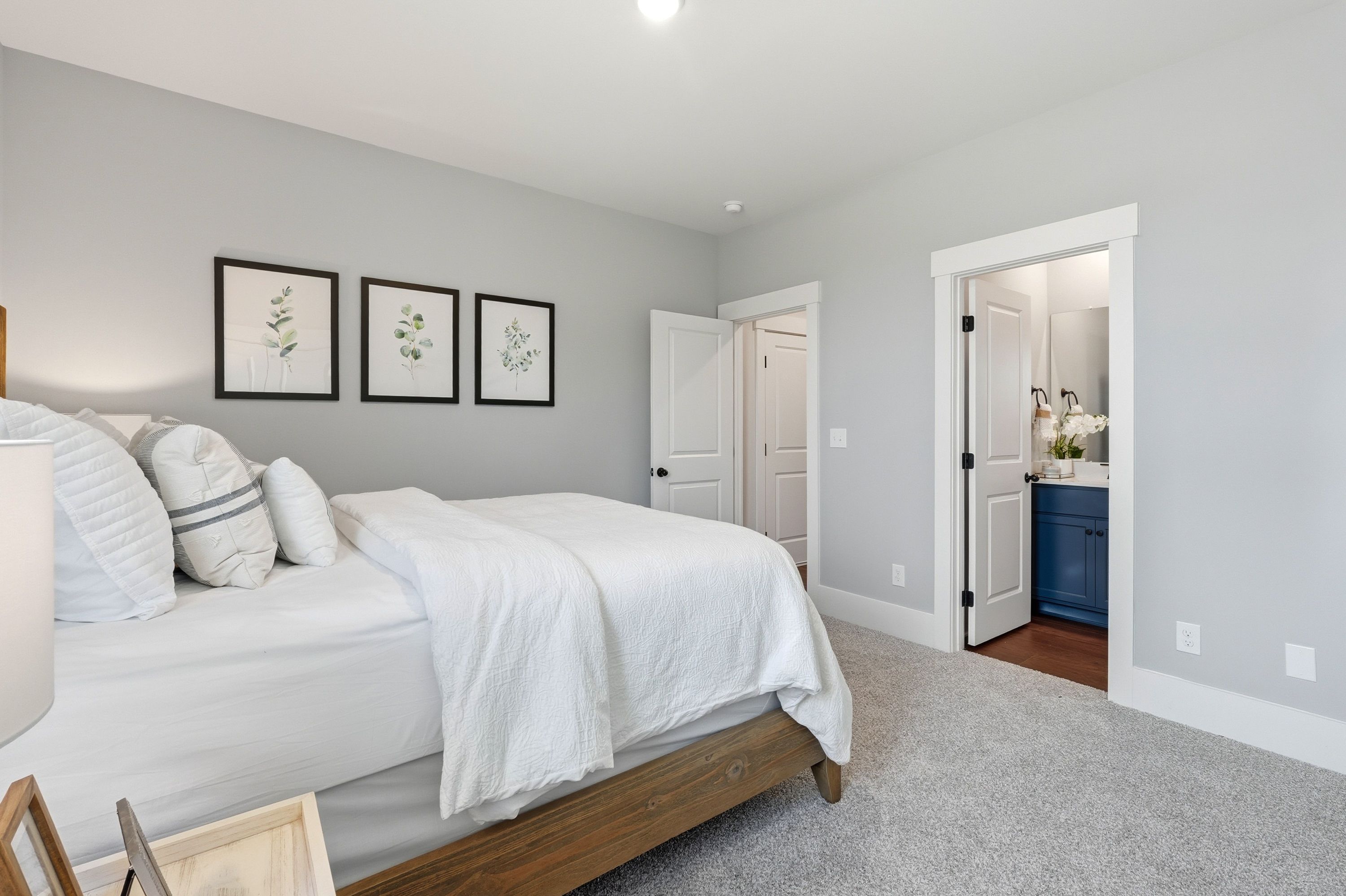 Spacious bedroom in Noble Ridge Cullman AL with gray walls, white king bed, botanical wall art, and blue vanity bathroom
