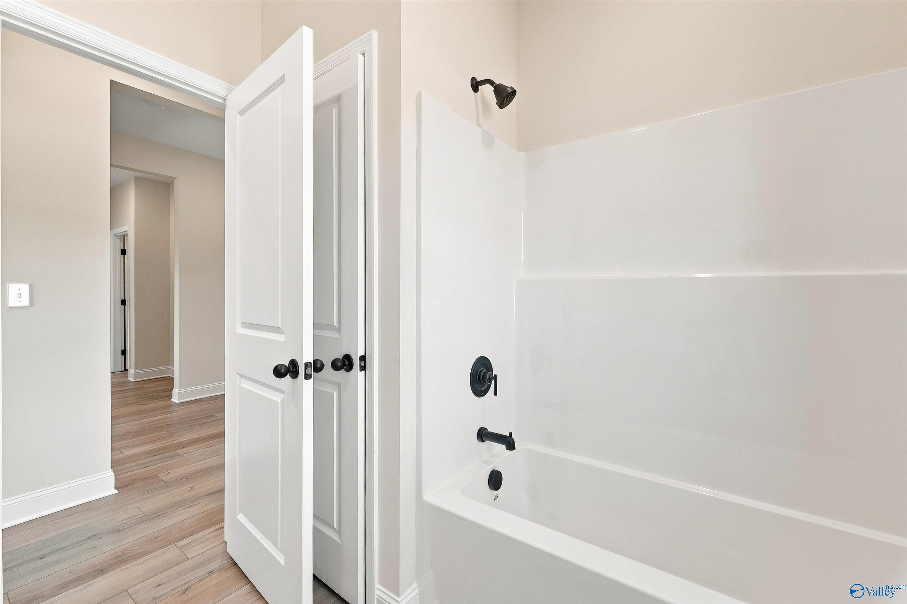 White bathtub with matte black fixtures beside open doors and hardwood flooring in Davidson Homes Everett B bathroom, Toney, Alabama