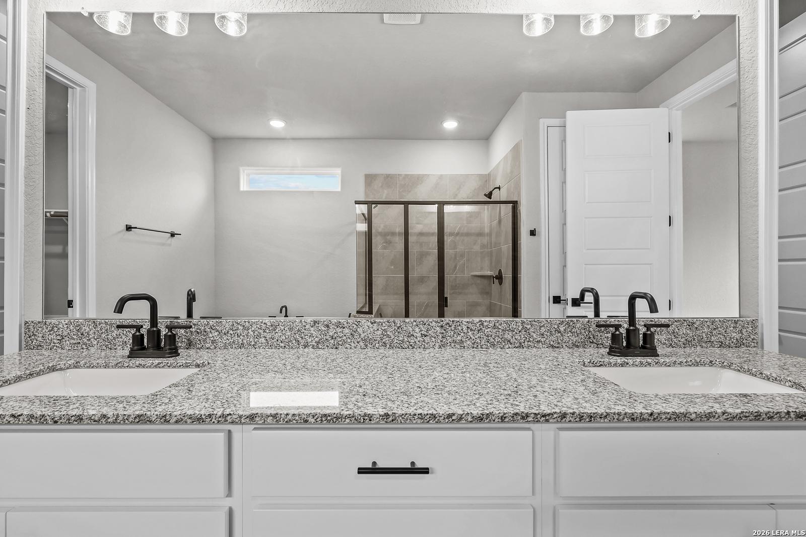 Modern master bathroom with double granite vanity, black faucets, frameless mirror, and glass shower in Davidson Homes The Belmont H, Castroville, Texas