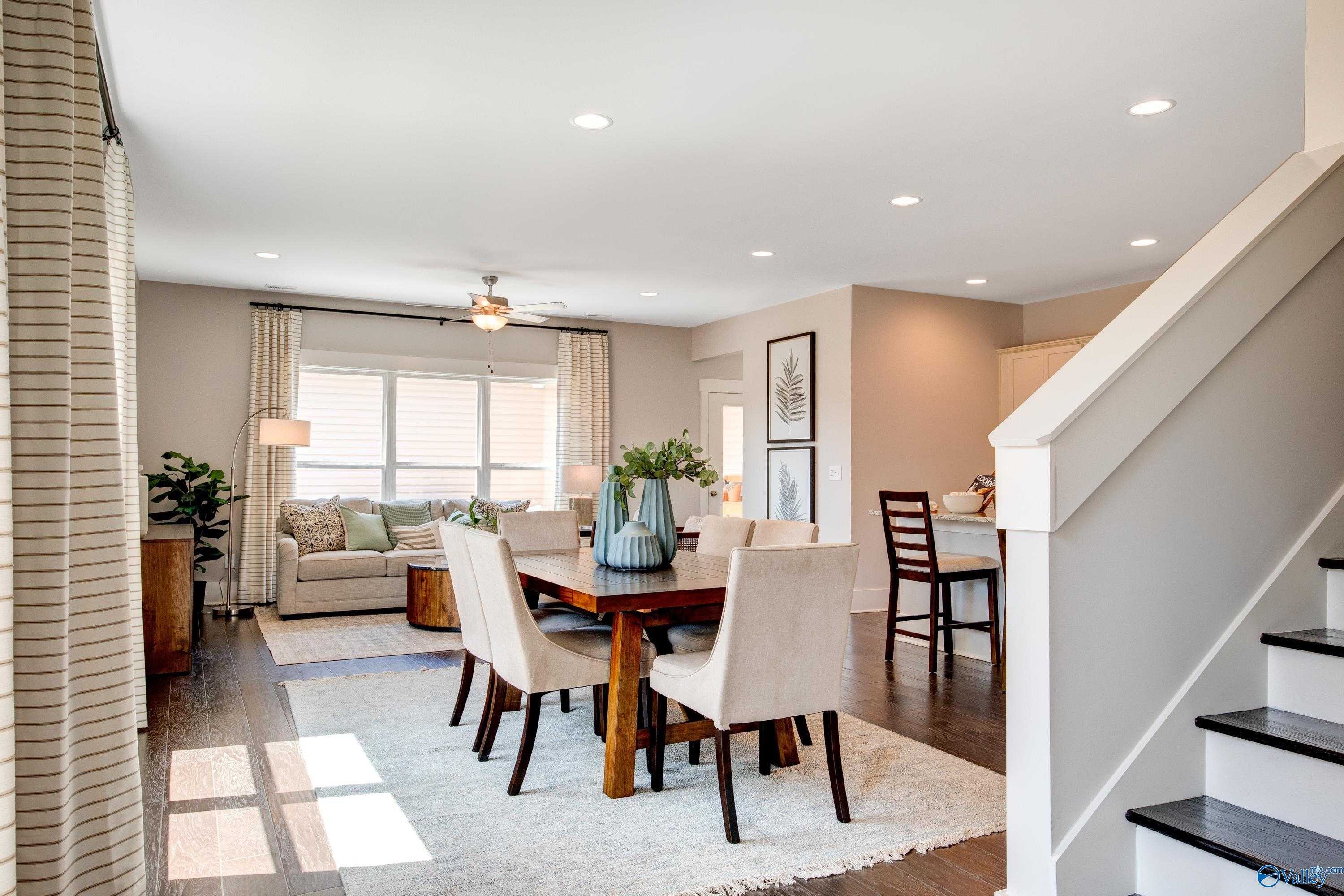 Bright open-concept dining-living area with wooden table, beige chairs, sofa, and staircase in Davidson Homes The Covington C, Decatur, Alabama