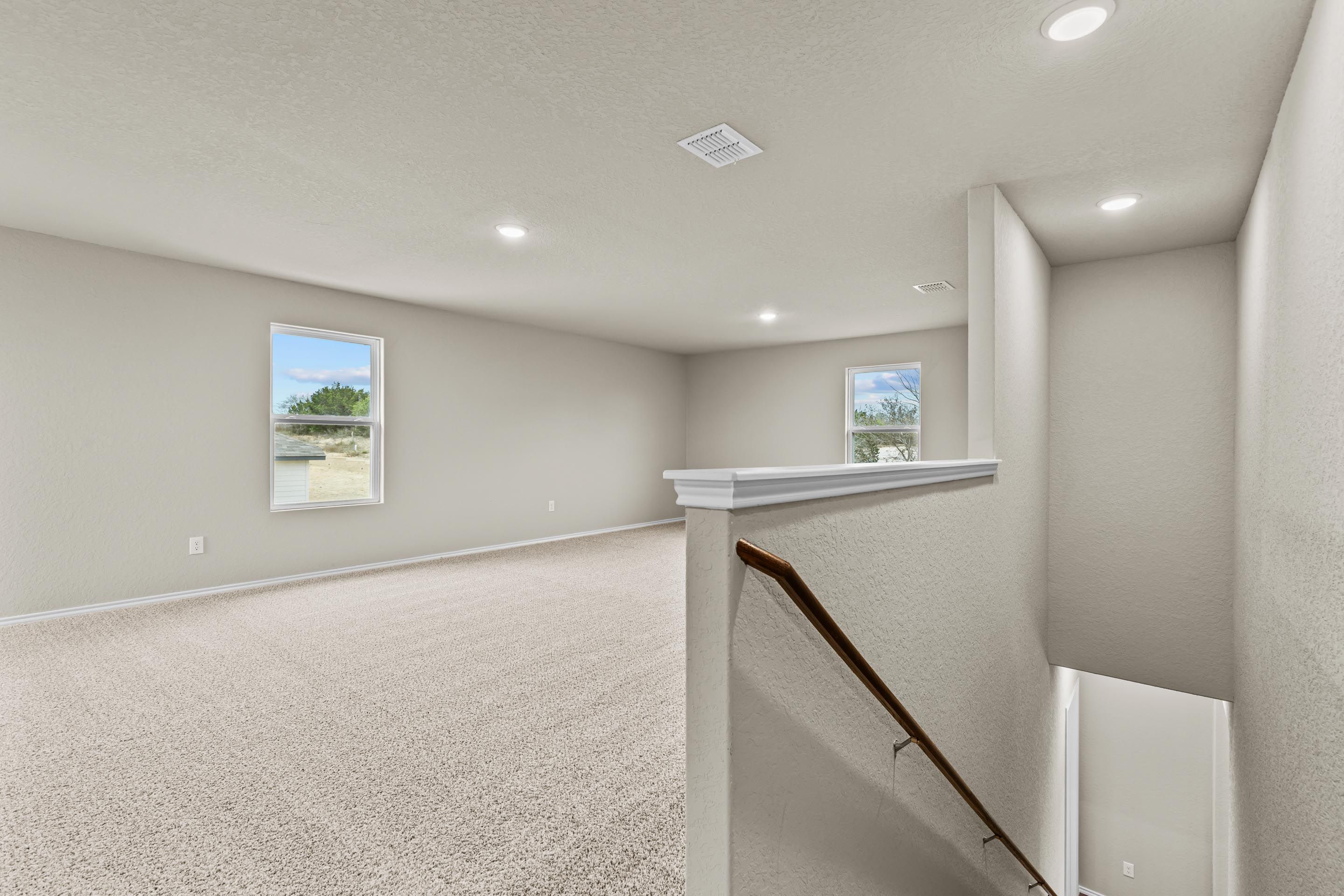 Spacious upstairs bonus room with beige walls, large windows, recessed lights, and wooden staircase railing in Davidson Homes The Douglas F, San Antonio