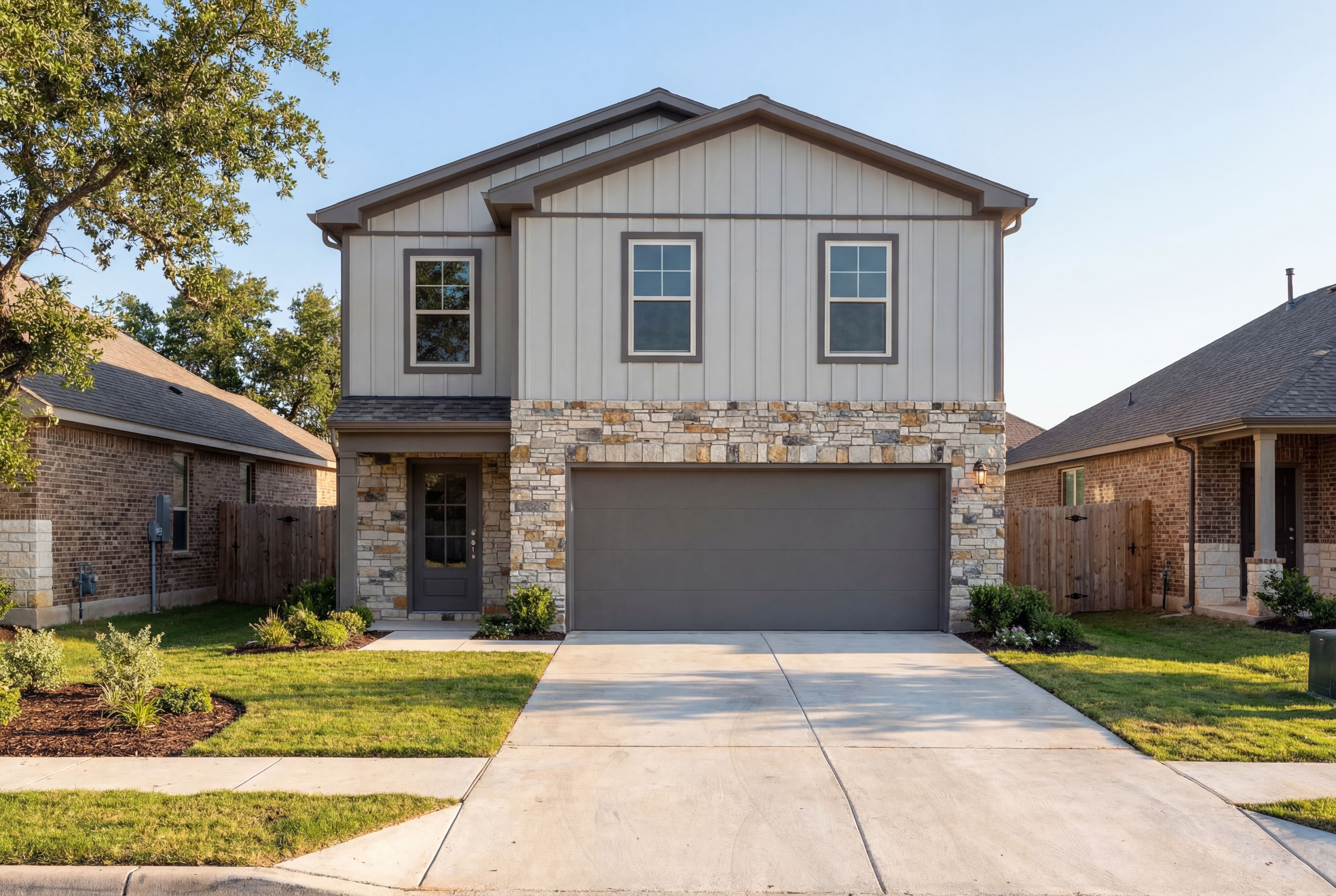 Two-story The Brazos E home elevation with gray board and batten siding, stone accents, two-car garage, and lush landscaped yard in Magnolia, Texas