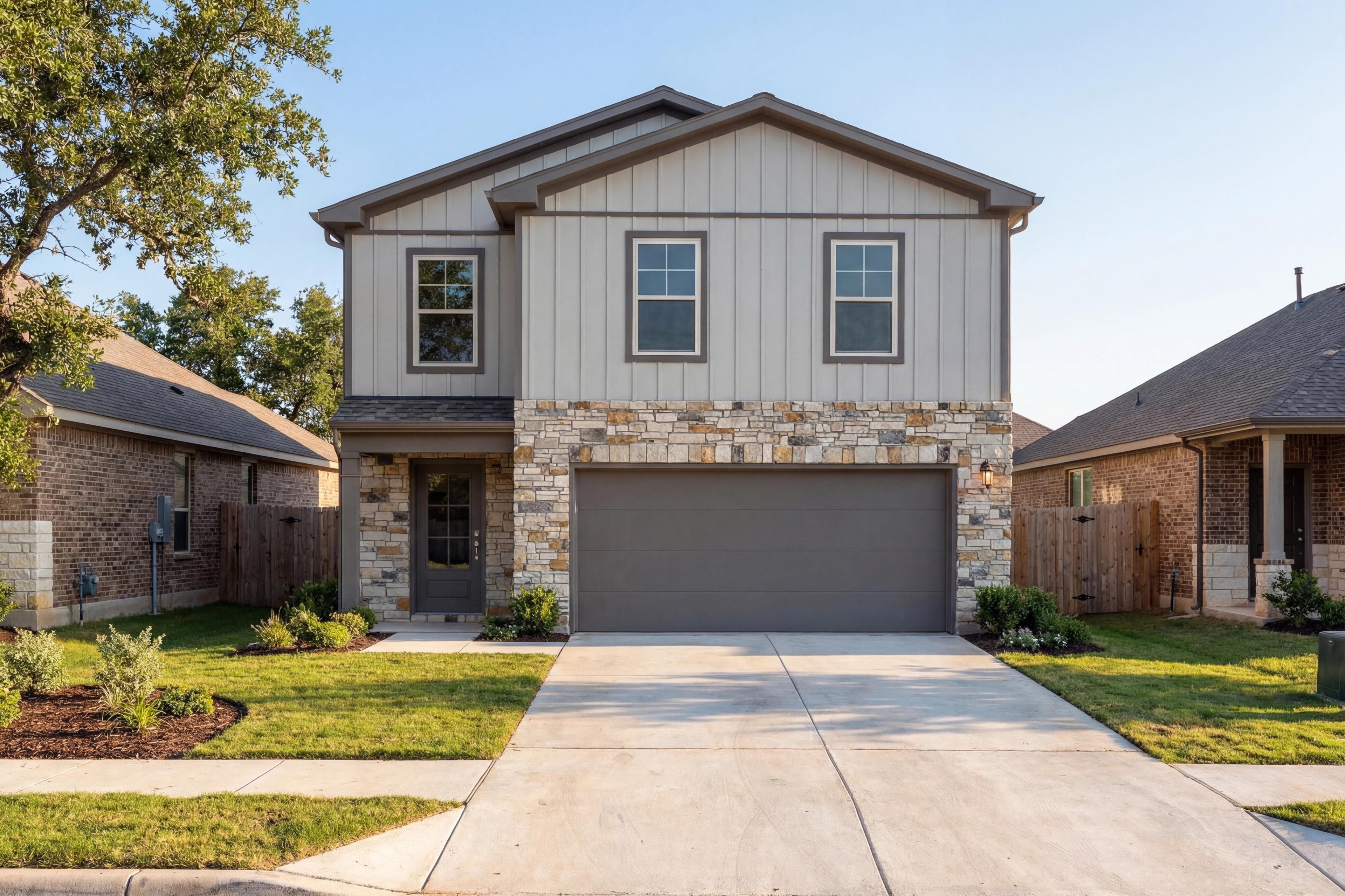 Two-story The Brazos E home elevation with gray board and batten siding, stone accents, two-car garage, and lush landscaped yard in Magnolia, Texas