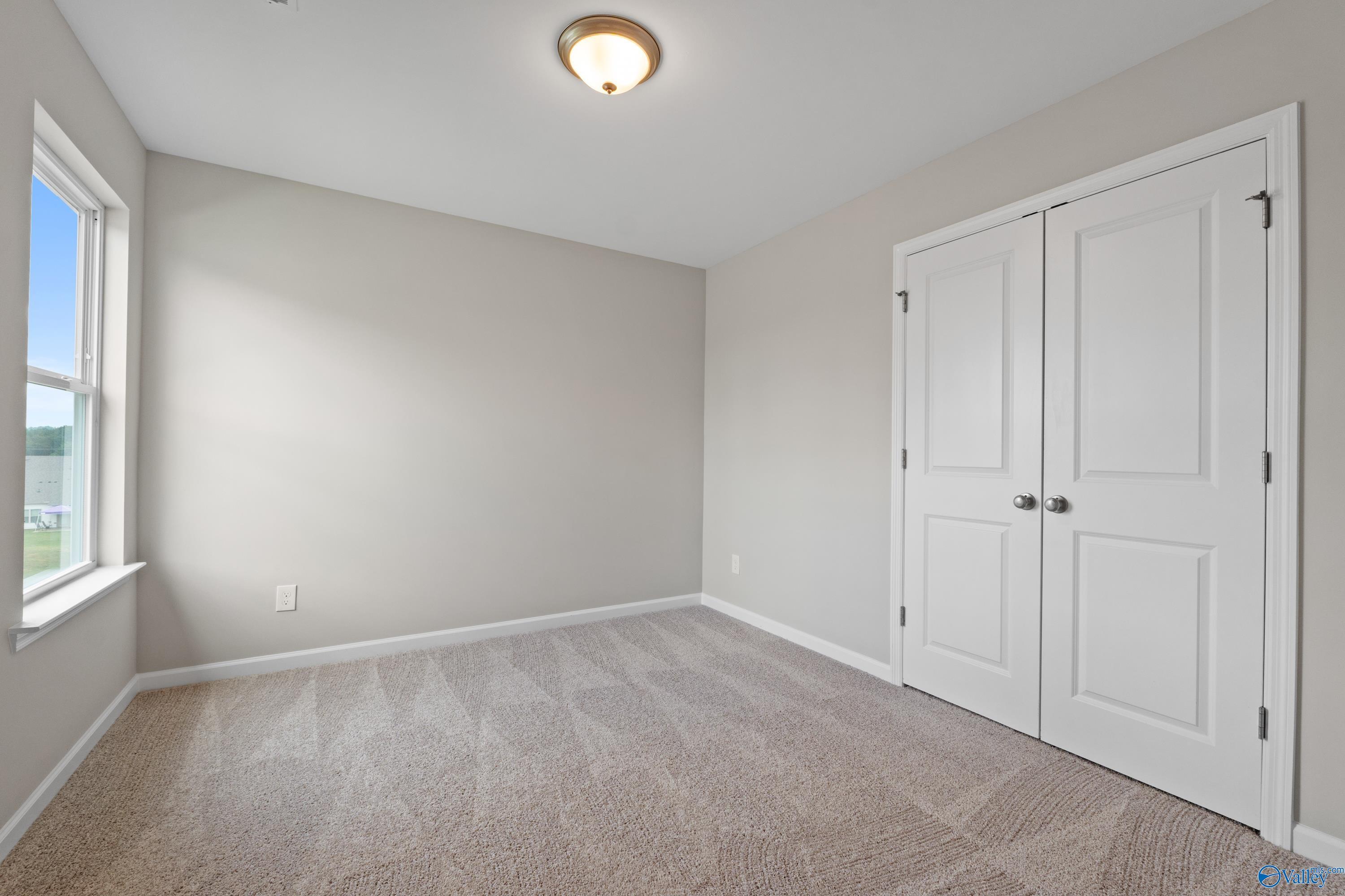 Empty secondary bedroom featuring light gray walls, beige carpet, large window, and double closet doors in Davidson Homes The Augusta, Madison AL