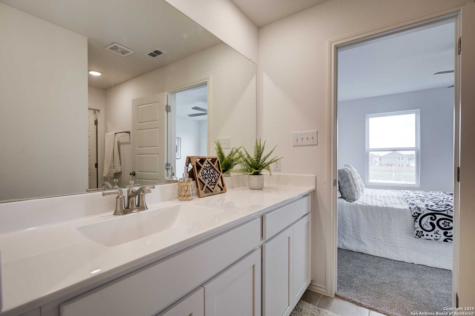Modern master bathroom with white double vanity, large mirror, plants, and adjacent bedroom in Davidson Homes The Trinity A, San Antonio