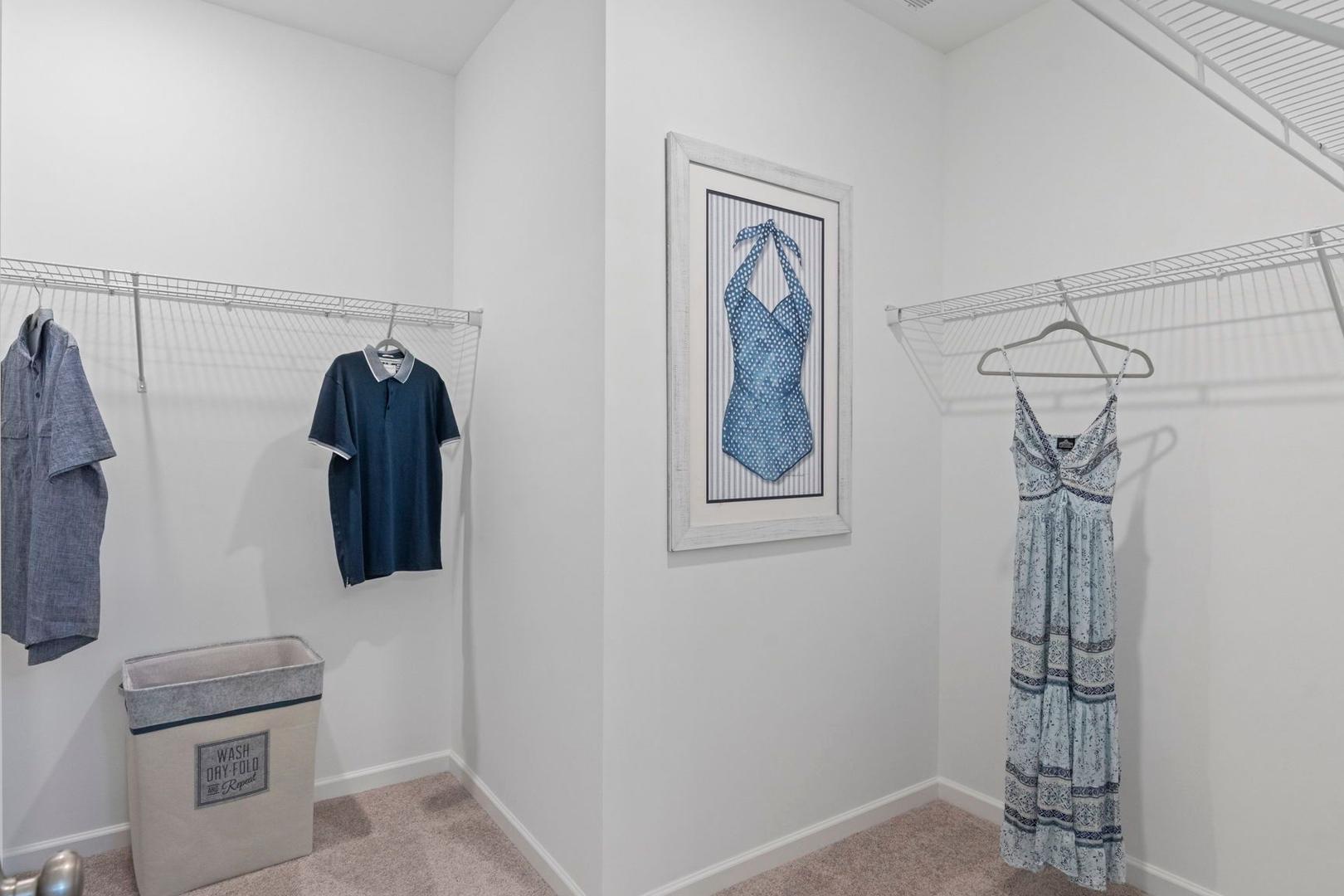 Spacious walk-in closet in The Phoenix A master suite with hanging shirts, dresses, framed blue gown art, and white hamper on beige carpet
