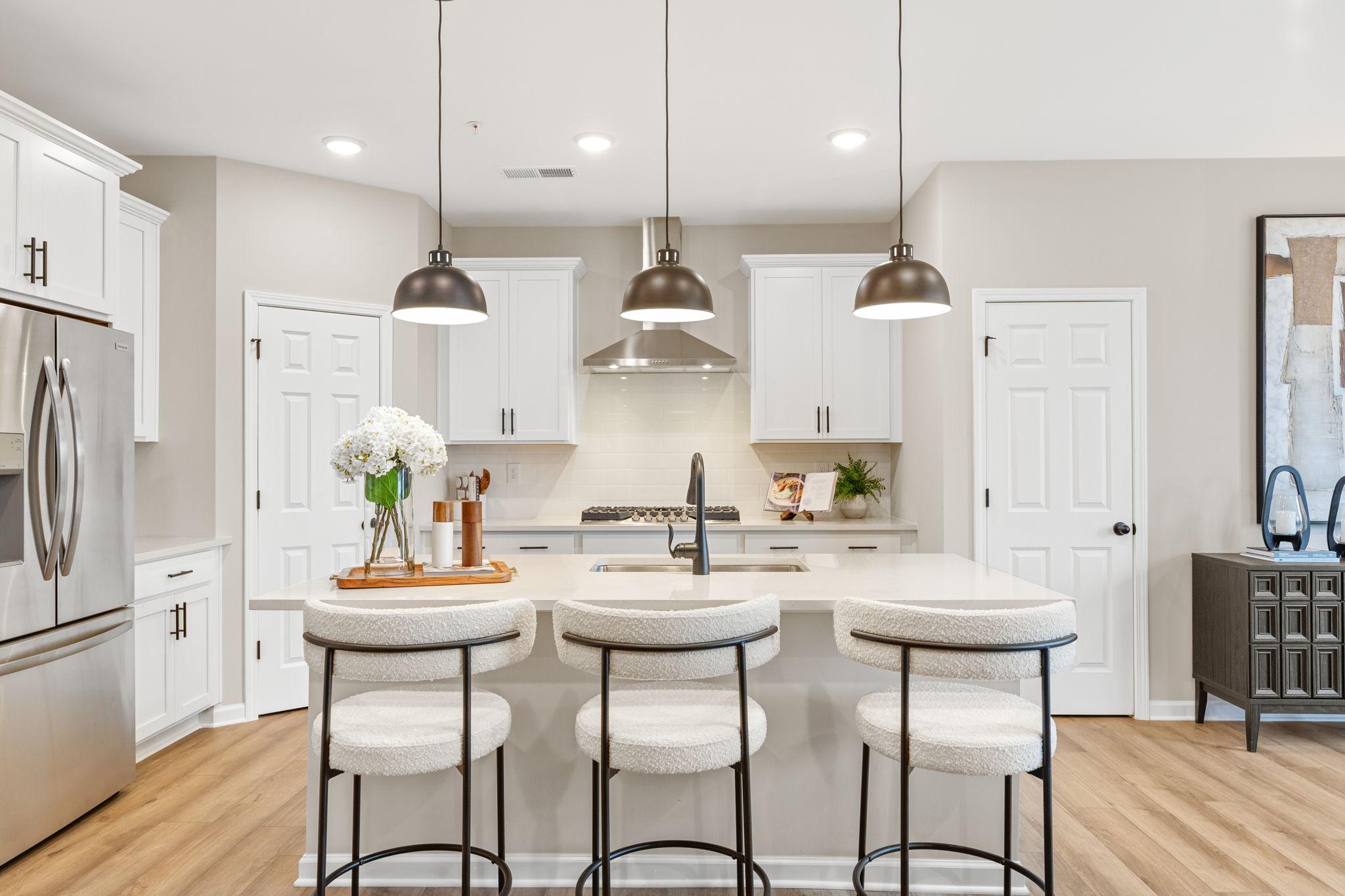 Modern white kitchen island with upholstered bar stools, stainless appliances, and pendant lights in The Durham B, Cumming, GA