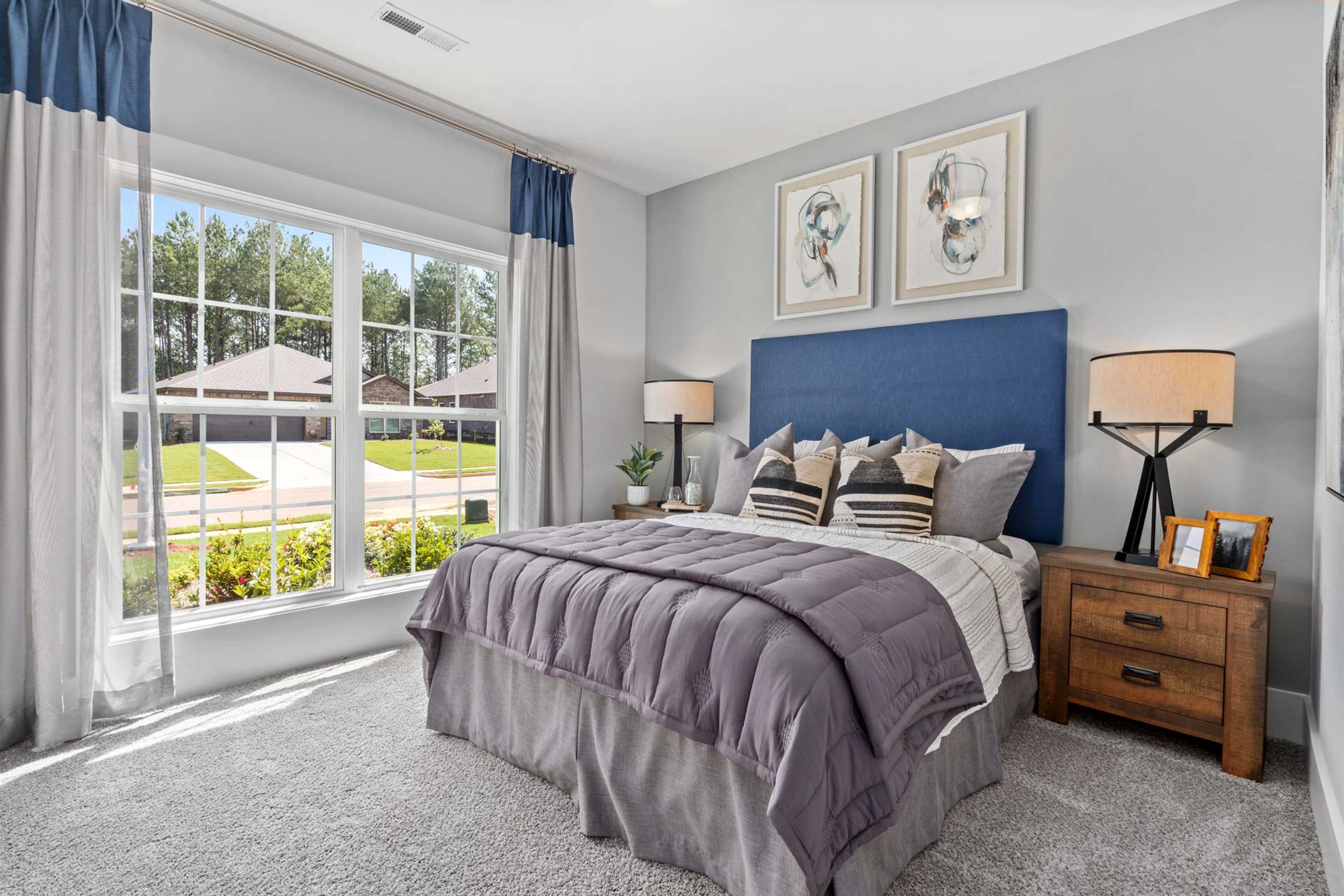 Spacious primary bedroom at Blue Spring in Huntsville Alabama with large windows overlooking neighborhood gray walls upholstered blue headboard plush purple bedding