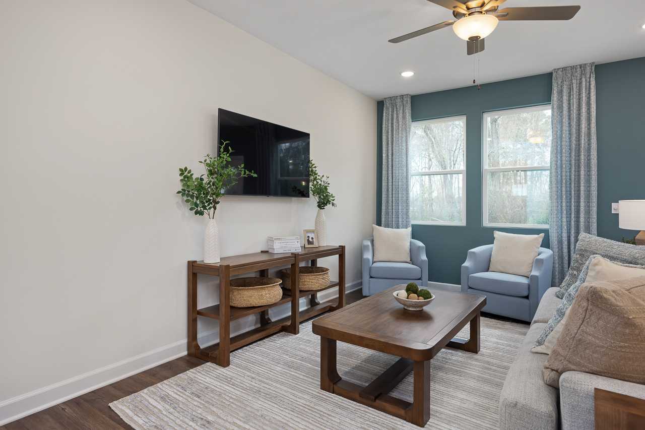 Cozy living room at Gregory Village Townhomes in Lillington NC with mounted TV, wooden console table, blue accent chairs, beige sofa, and large curtained windows