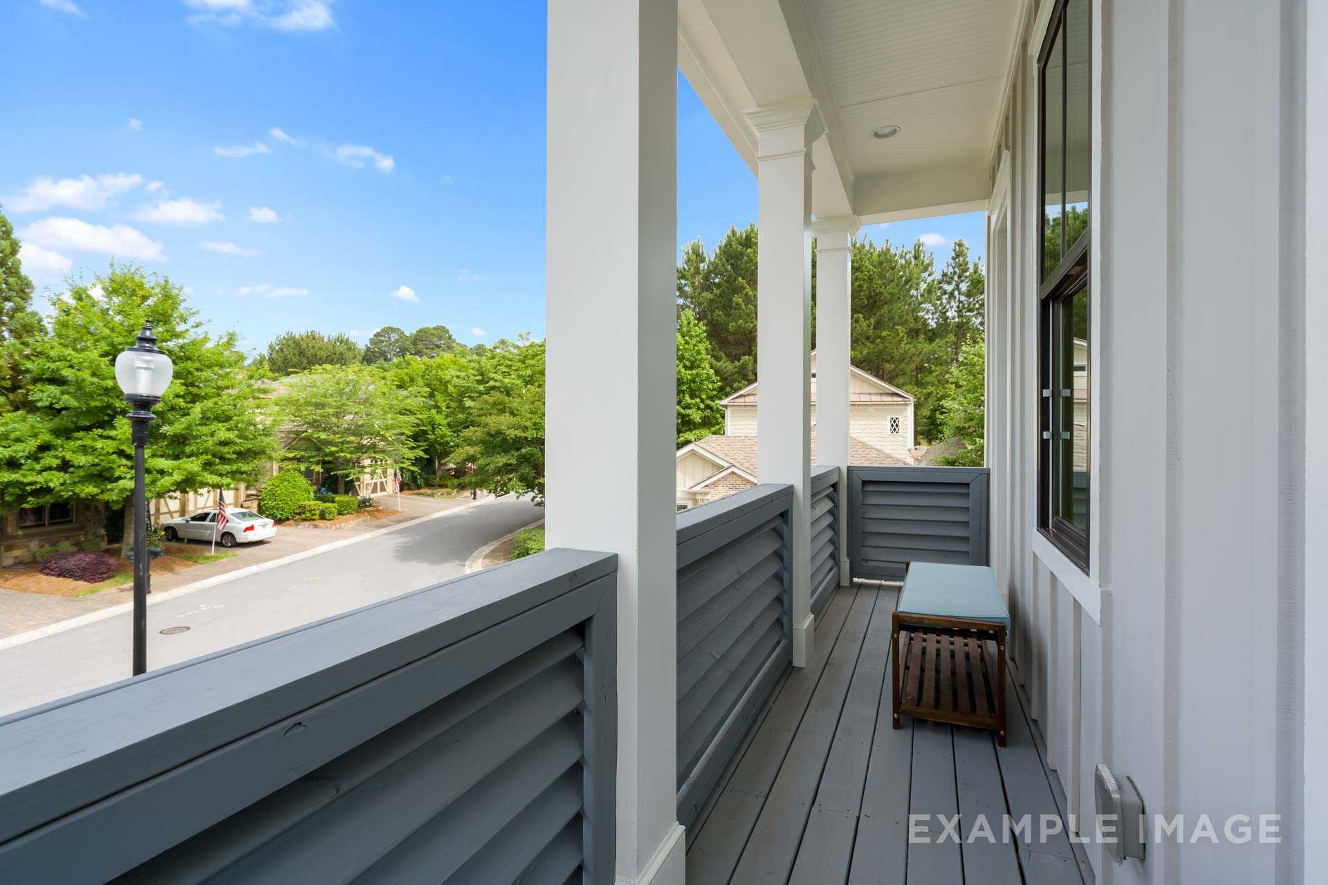 Spacious upper-floor balcony of The Seaside home design with white columns, wooden bench, and wooded neighborhood view in Woodstock GA