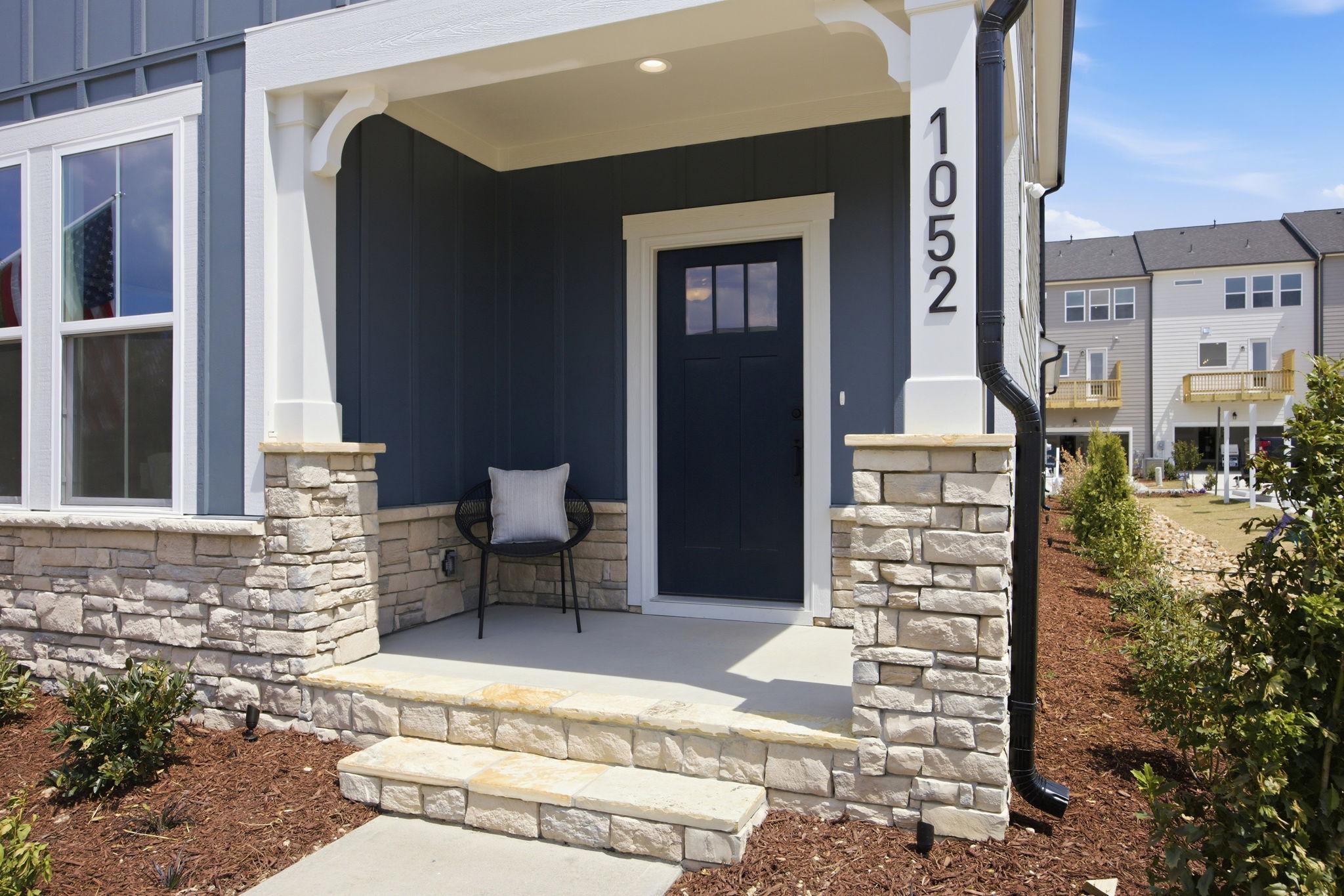 Covered front porch of The Burke E by Davidson Homes featuring stone accents, blue siding, and a modern entryway.