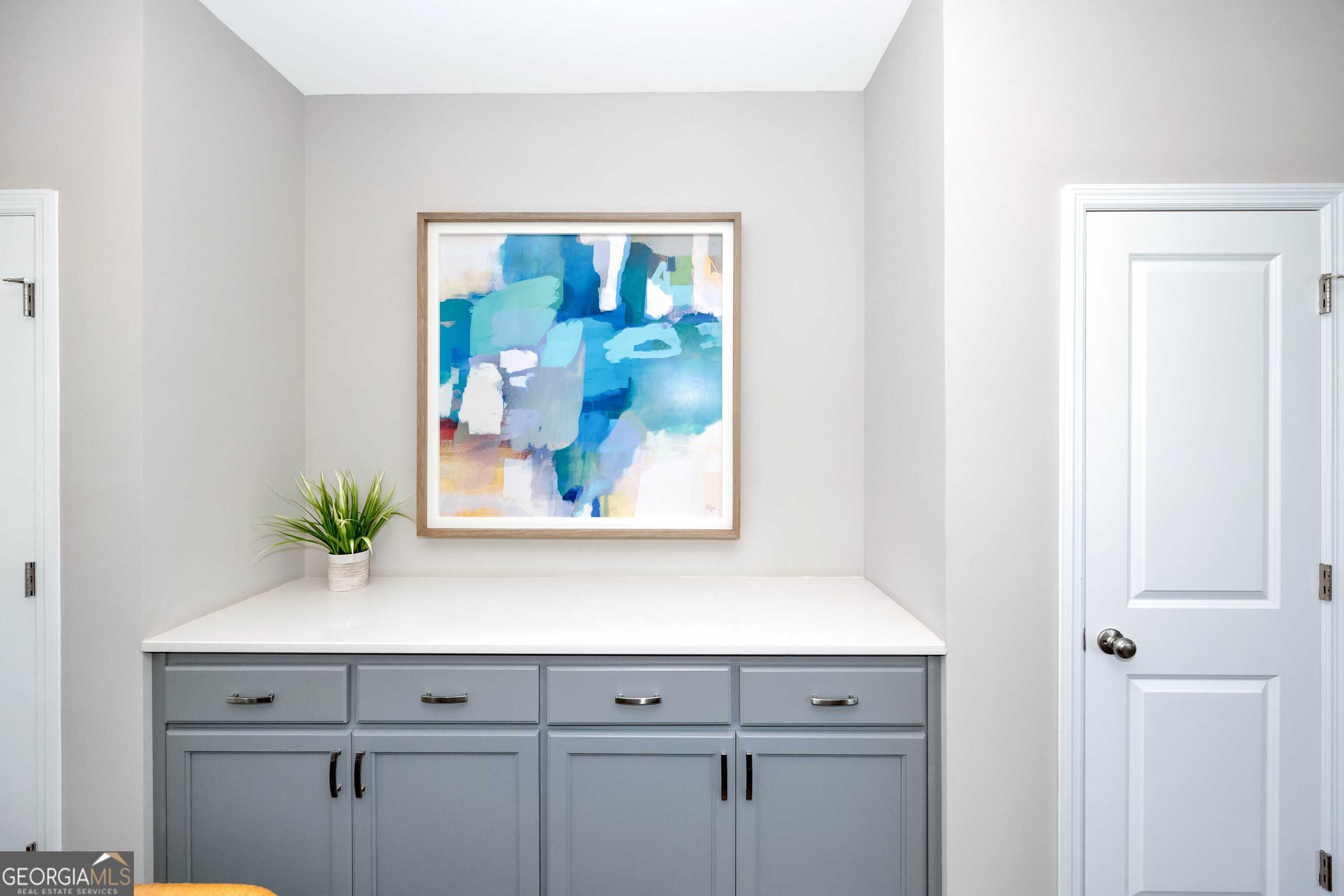 Modern laundry room featuring gray cabinets, white countertop, abstract blue artwork, and potted plant in Evermore Homes The Stella, Perry, GA
