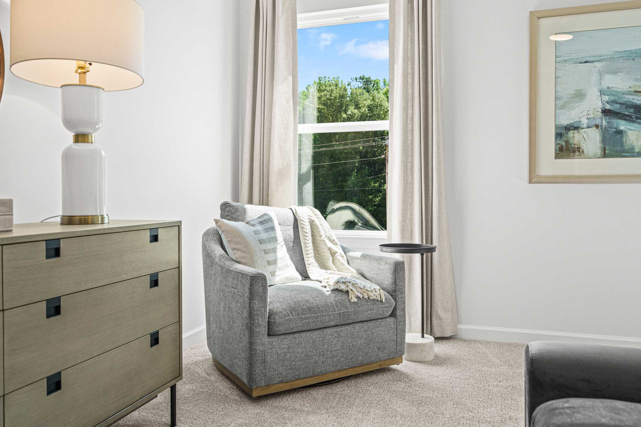 Cozy gray armchair with pillows and throw in Cedar Farms Winder Georgia home, beside lamp, dresser, and tree-view window