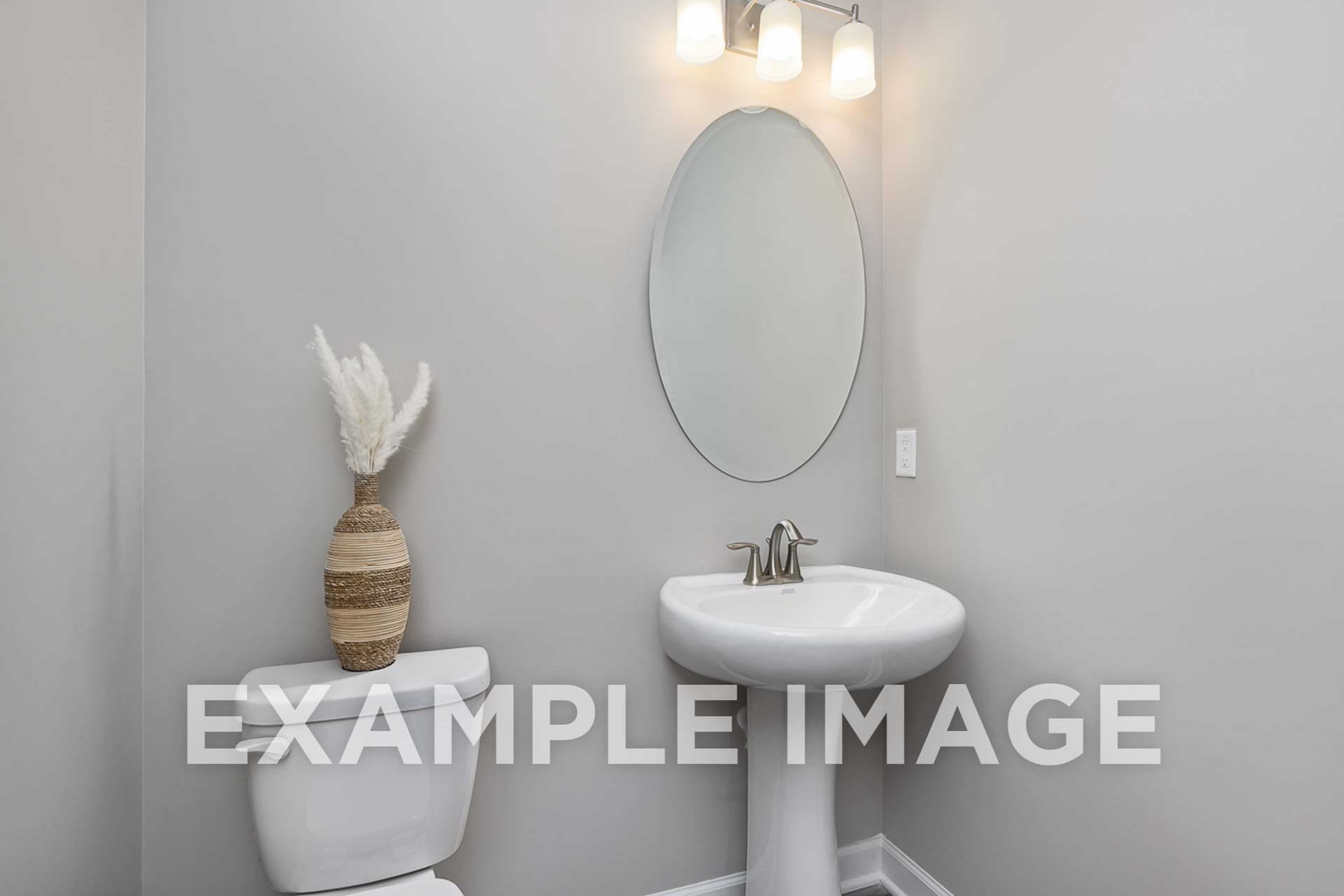 The Ash powder room with oval mirror above pedestal sink, white toilet, potted feather plant, gray walls, soft lighting