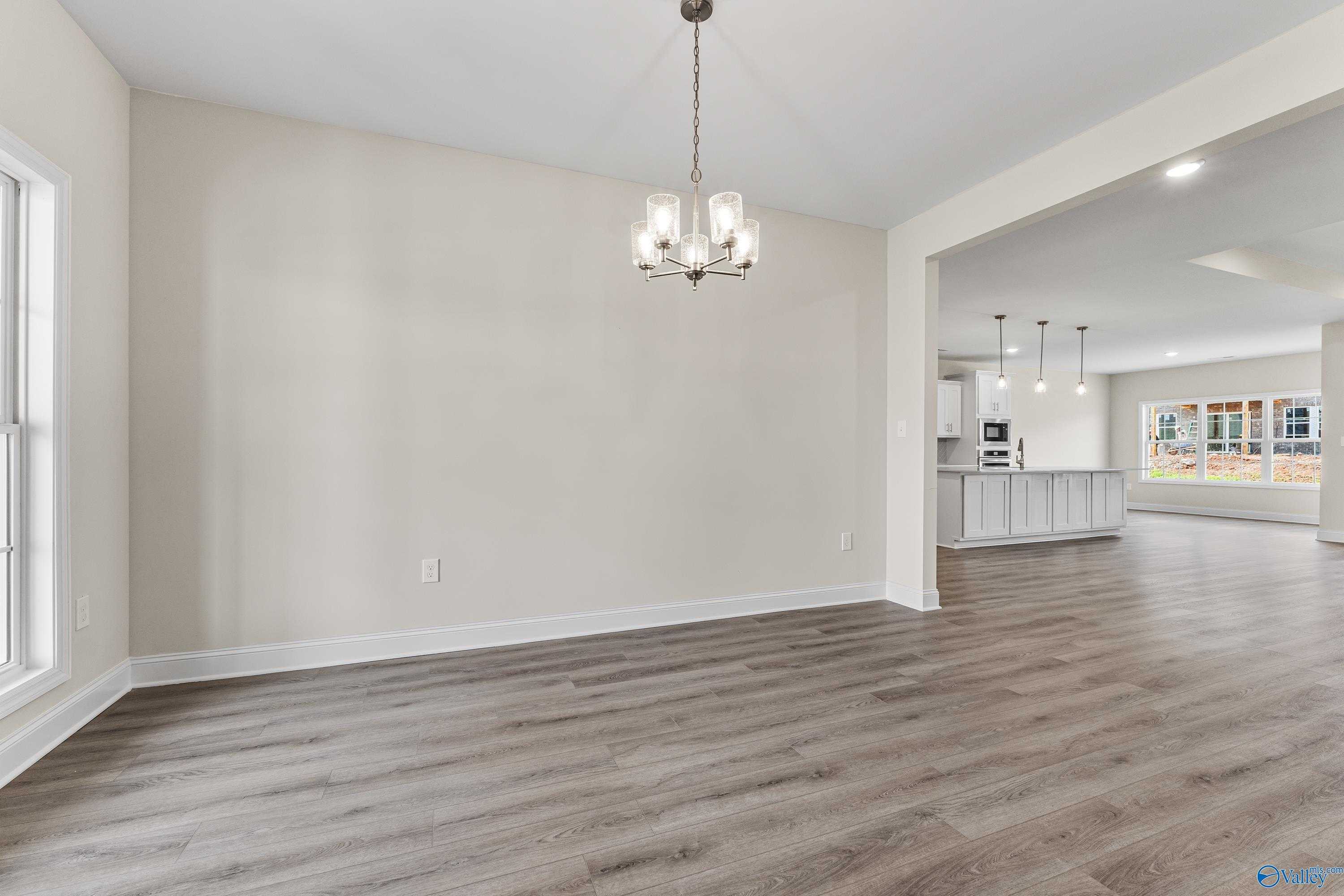 Spacious dining room with chandelier, beige walls, and hardwood floors open to white kitchen cabinets in The Finleigh, Toney, Alabama