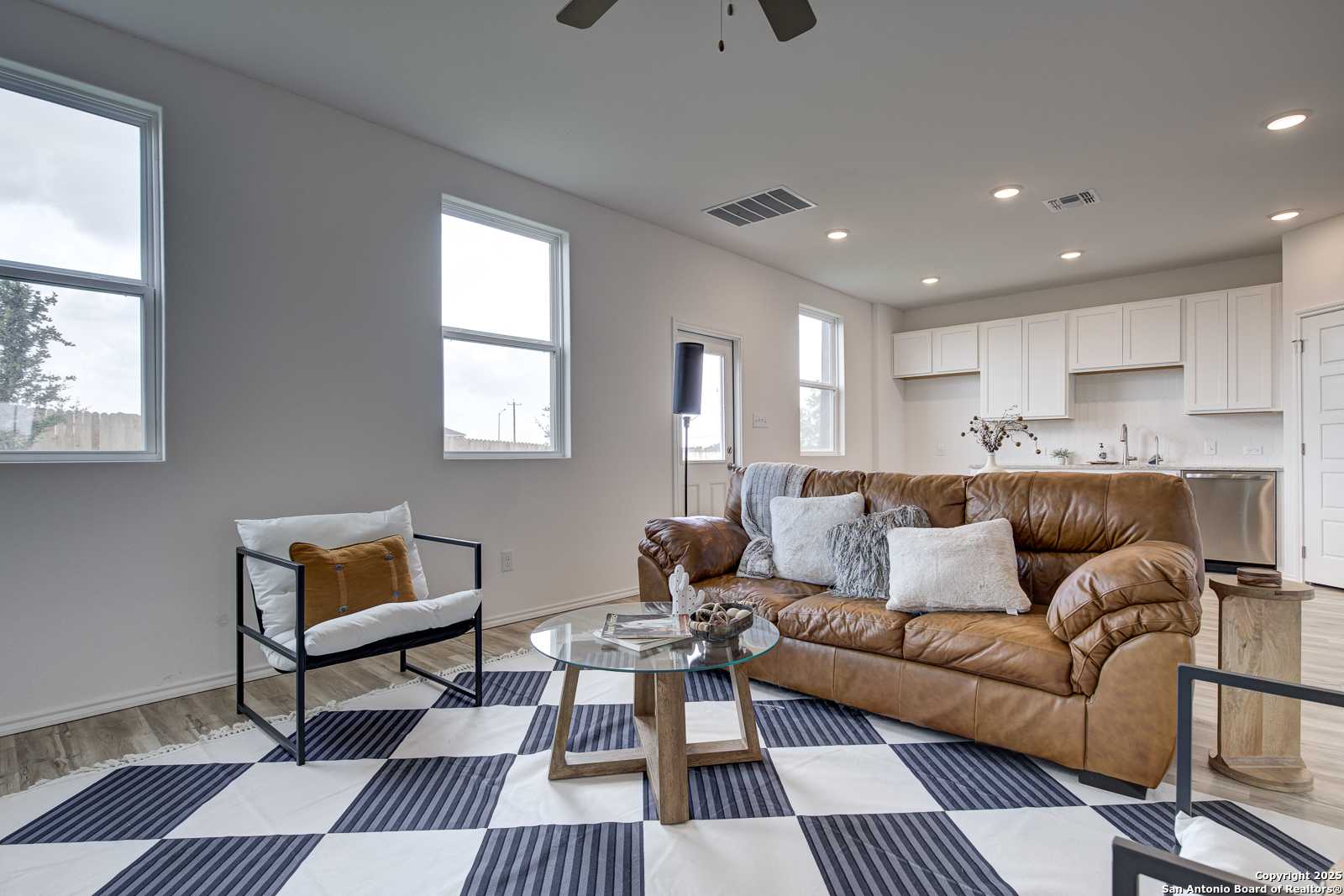 Cozy living room with brown leather sofa, white armchair, and checkered rug opening to white kitchen cabinets in Davidson Homes The Trinity A, San Antonio
