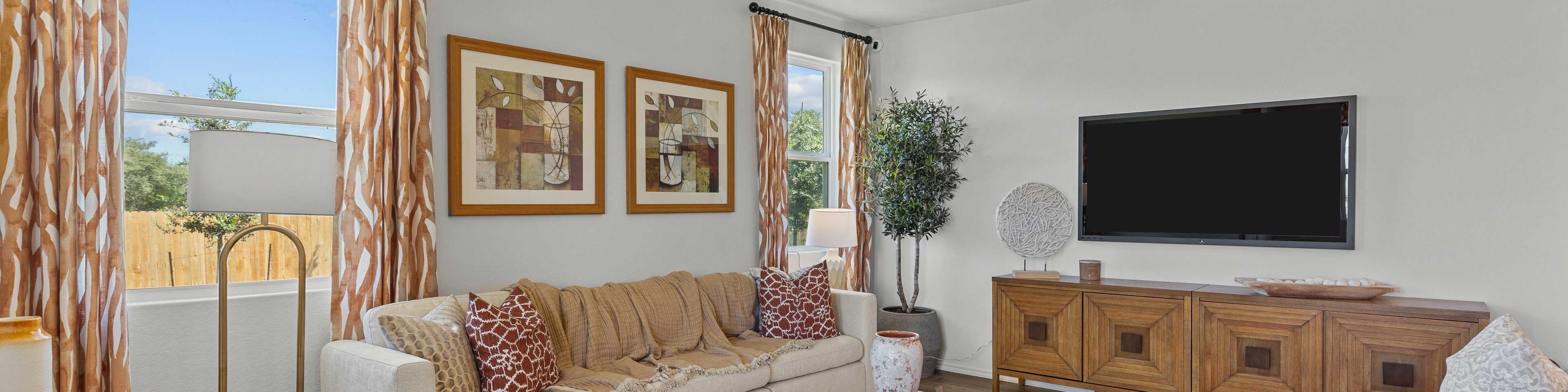 Cozy living room at Royal Crest in San Antonio Texas featuring beige sofa, wooden media console, TV, and large windows with patterned curtains
