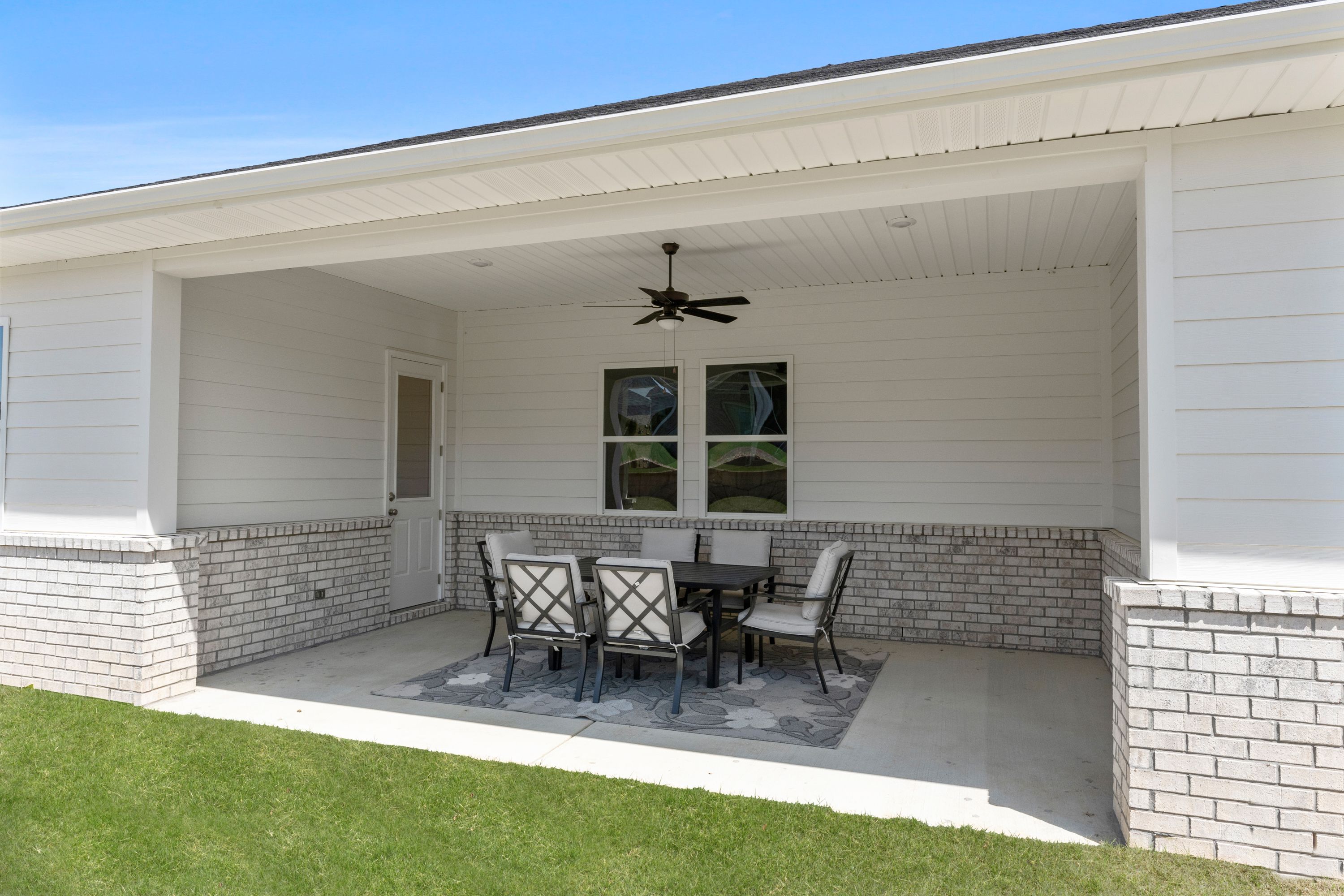 Spacious covered patio of The Haven D showcasing white brick exterior, ceiling fan, and elegant wicker dining set on gray rug