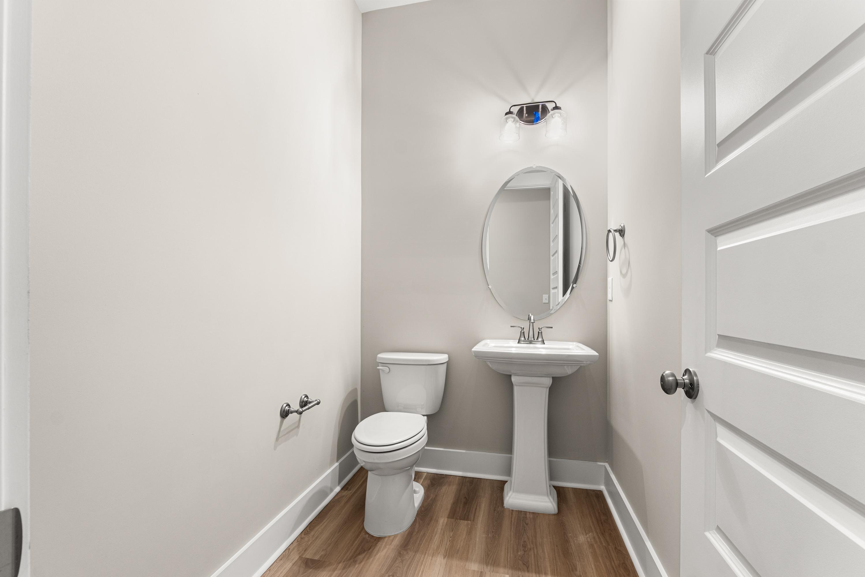 Cozy powder room in The Oxford C home design with white pedestal sink, round mirror, toilet, and light gray walls
