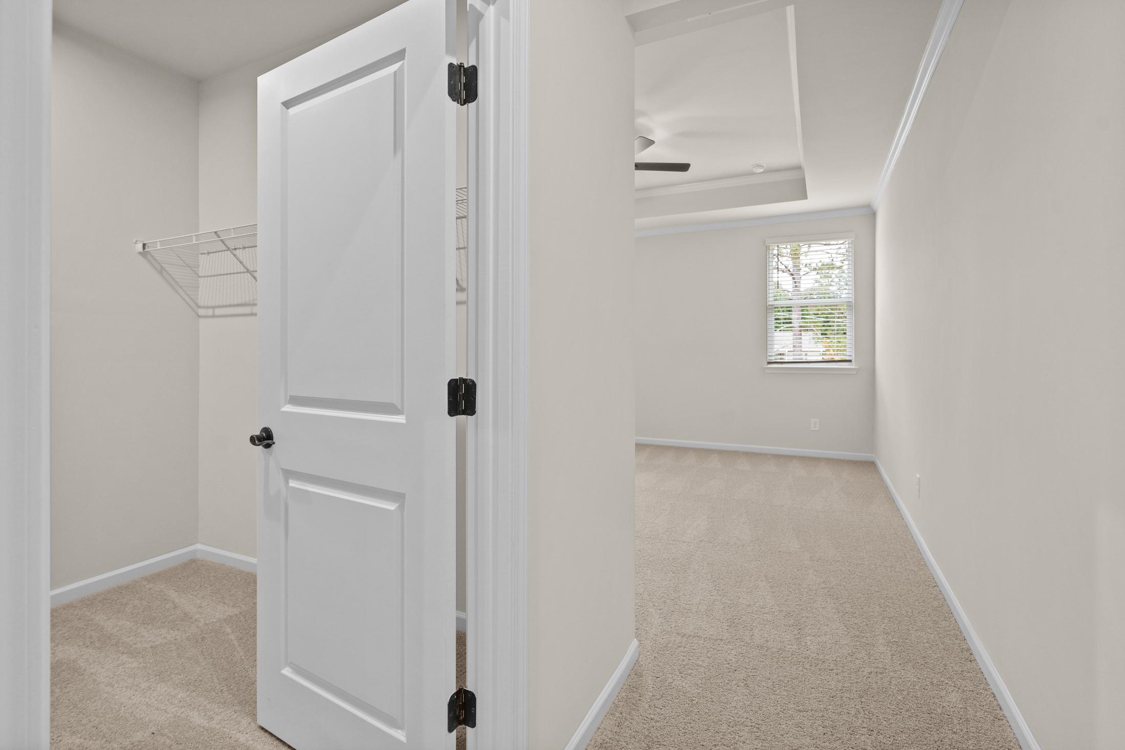 Spacious walk-in closet in The Glenwood with open white door, beige walls, carpeted floor, metal rod, and adjacent bedroom window