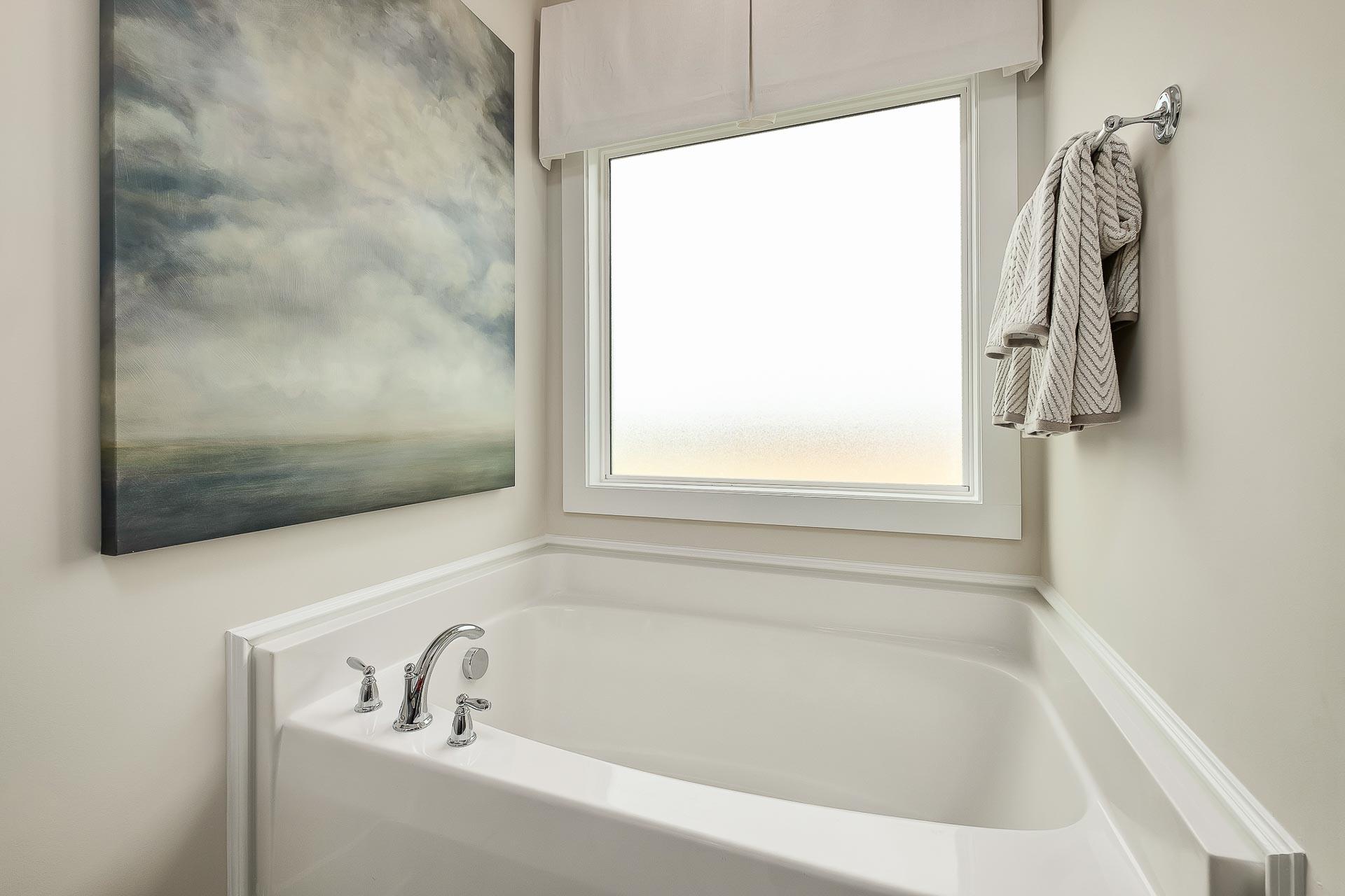 Spacious master bathroom in The Everett with white corner bathtub, chrome faucets, large window, and abstract seascape painting