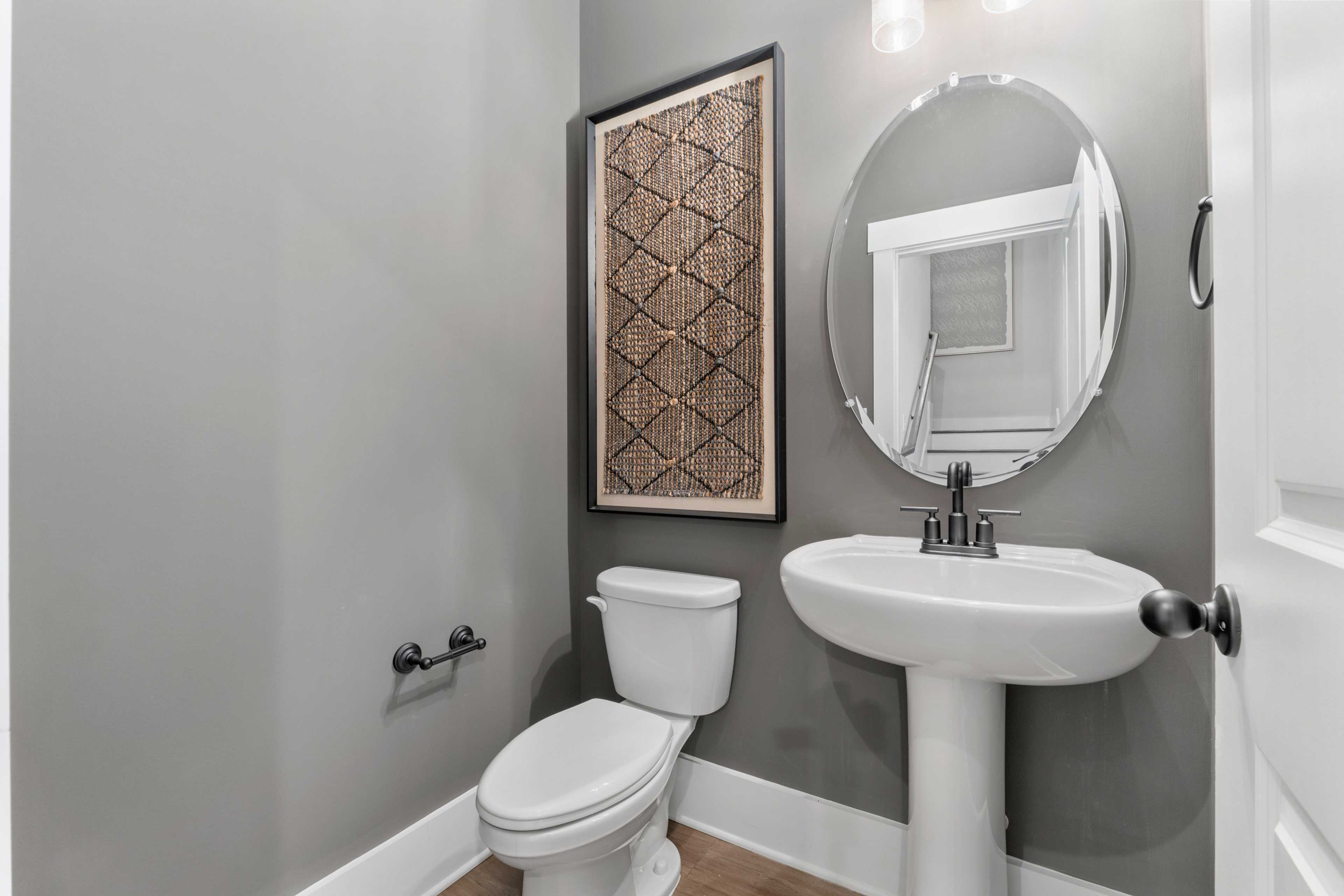 Modern powder room at Kendall Farms in Toney, Alabama with gray walls, woven wall art, round mirror, pedestal sink, and toilet