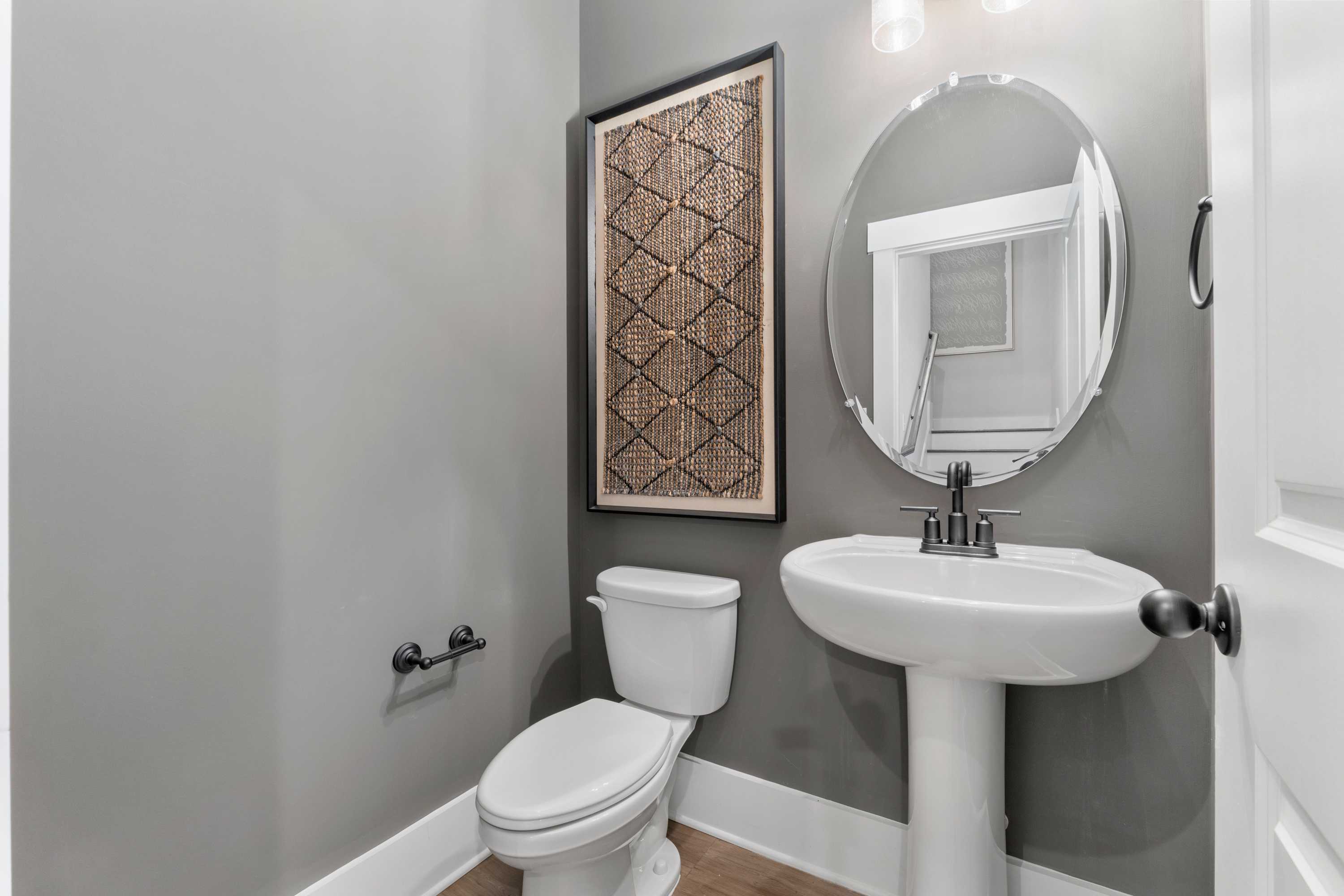 Modern powder room at Kendall Farms in Toney, Alabama with gray walls, woven wall art, round mirror, pedestal sink, and toilet