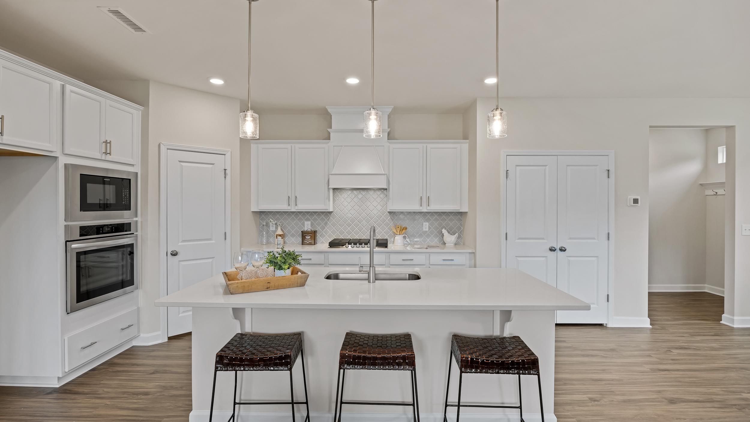 Spacious white kitchen in Gregory Village Lillington NC with central island, bar stools, pendant lights and hardwood floors by Davidson Homes