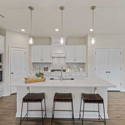 Spacious white kitchen in Gregory Village Lillington NC with central island, bar stools, pendant lights and hardwood floors by Davidson Homes