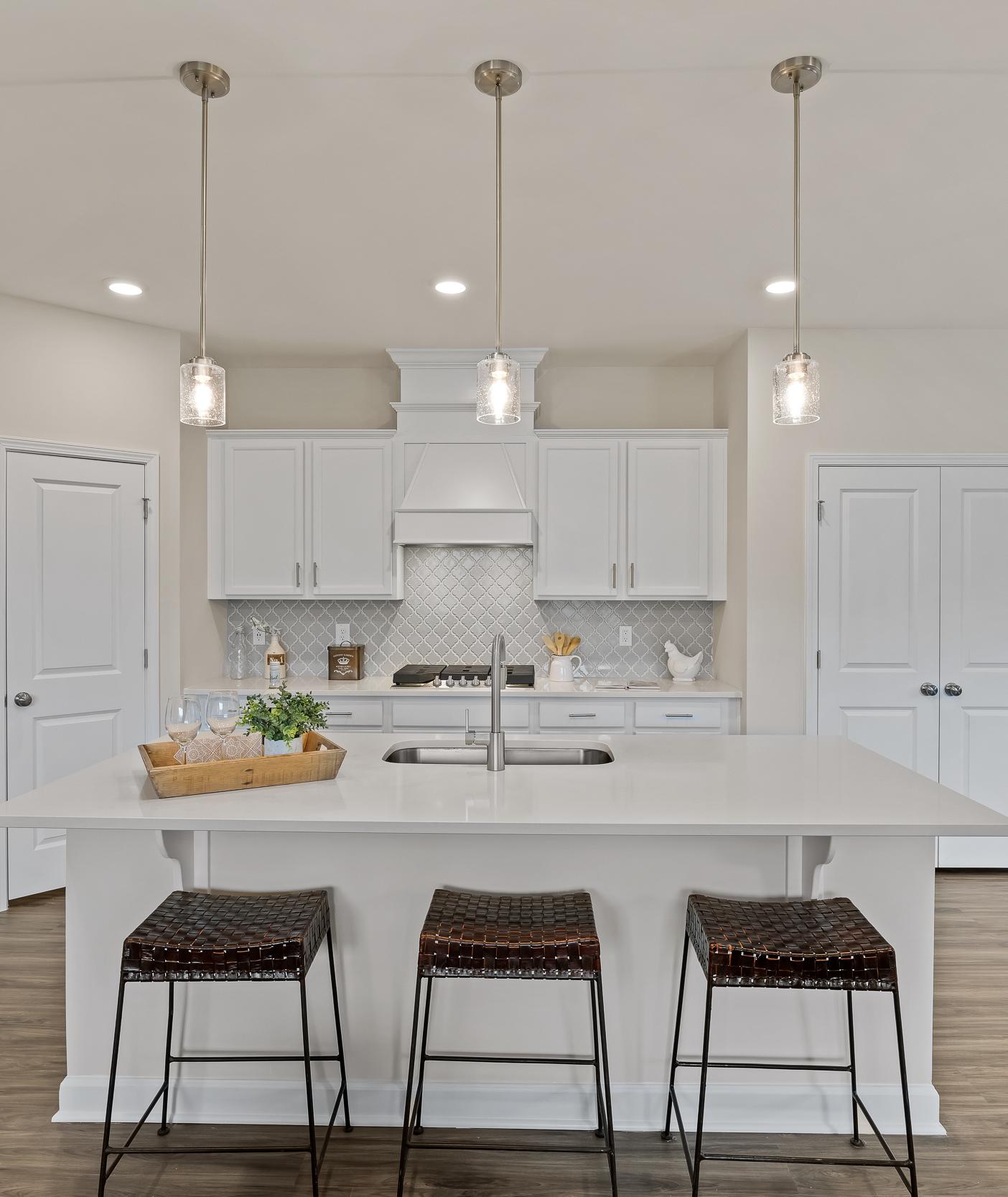 Spacious white kitchen in Gregory Village Lillington NC with central island, bar stools, pendant lights and hardwood floors by Davidson Homes
