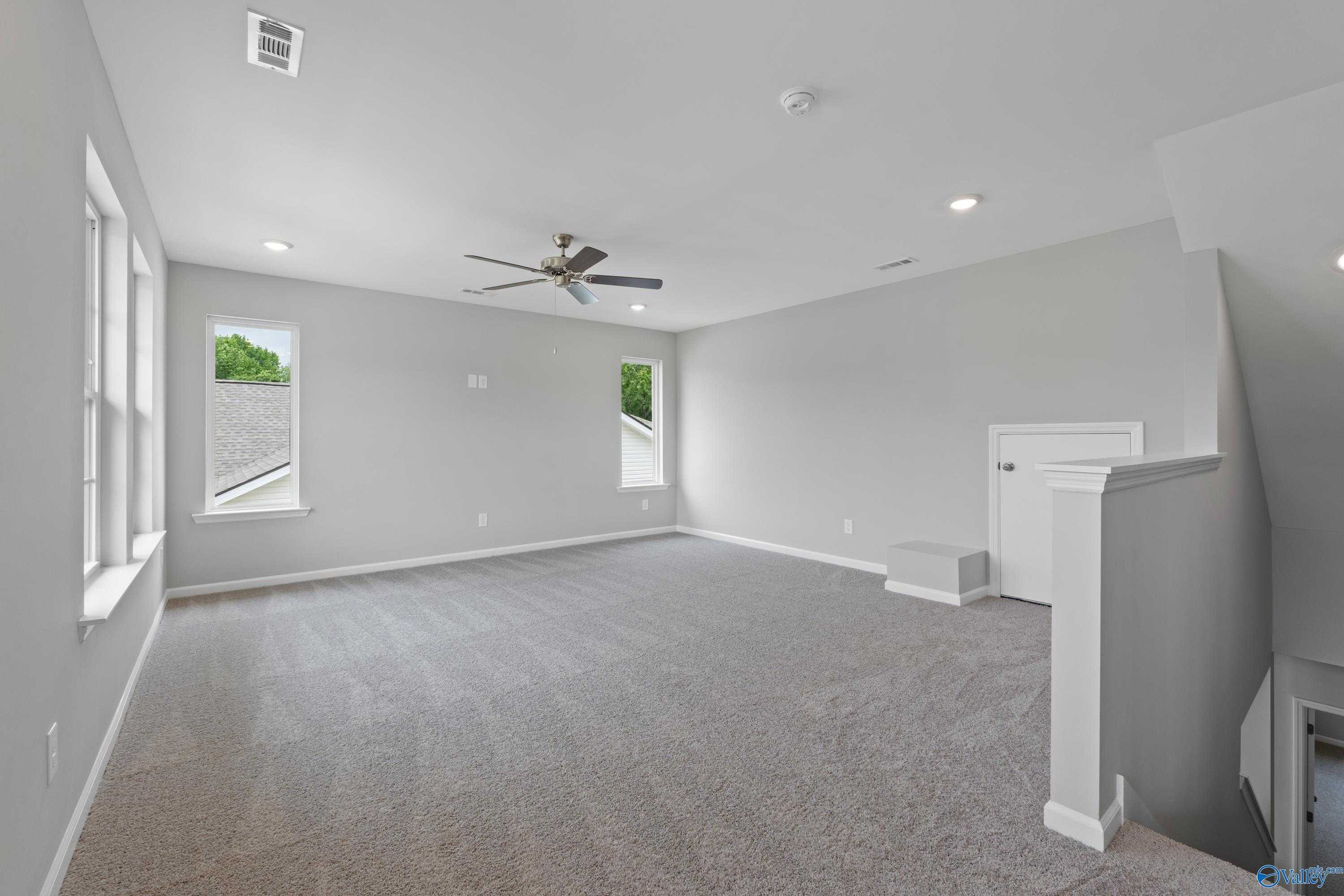 Spacious upstairs bonus room with gray carpet, ceiling fan, large windows overlooking trees, and overlook railing in Davidson Homes The Stella, Hazel Green, AL