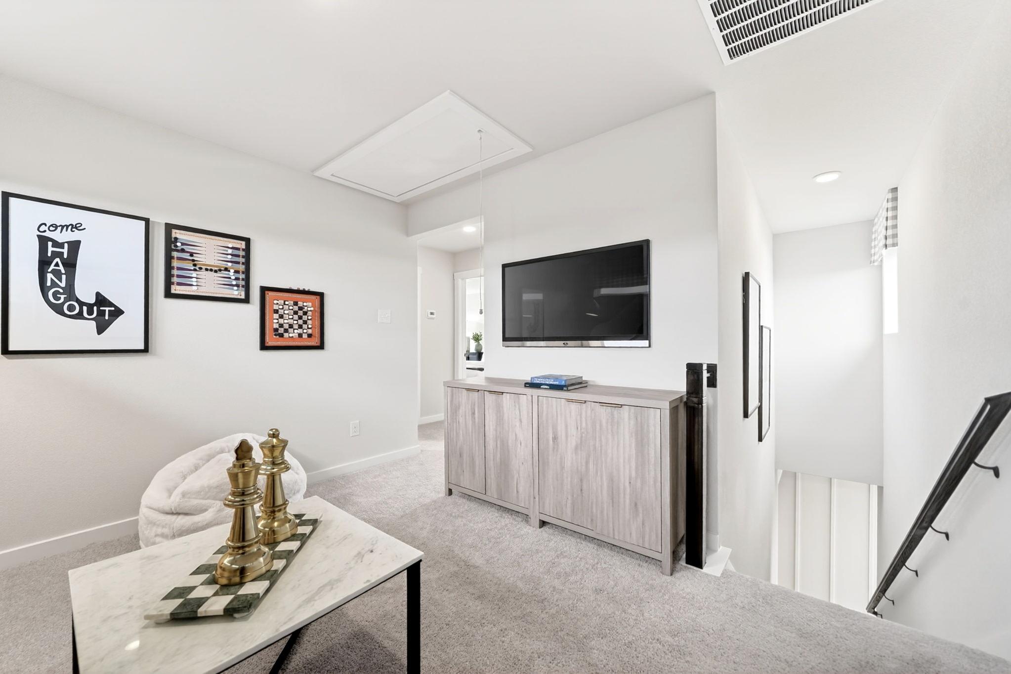 Cozy loft space at Heartland Texas by Davidson Homes with mounted TV, brass chess set on marble table, wall art, and gray carpet