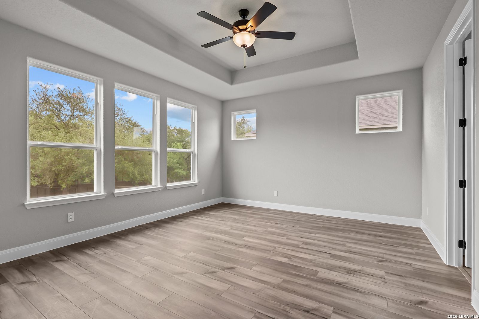 Bright bedroom with large windows overlooking trees, gray walls, ceiling fan, and hardwood floors in Davidson Homes The Belmont H, Castroville Texas