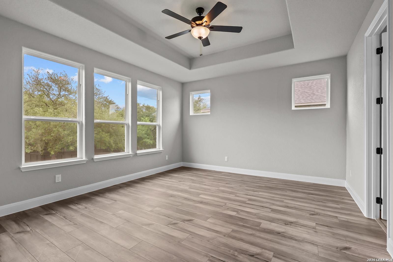 Bright bedroom with large windows overlooking trees, gray walls, ceiling fan, and hardwood floors in Davidson Homes The Belmont H, Castroville Texas