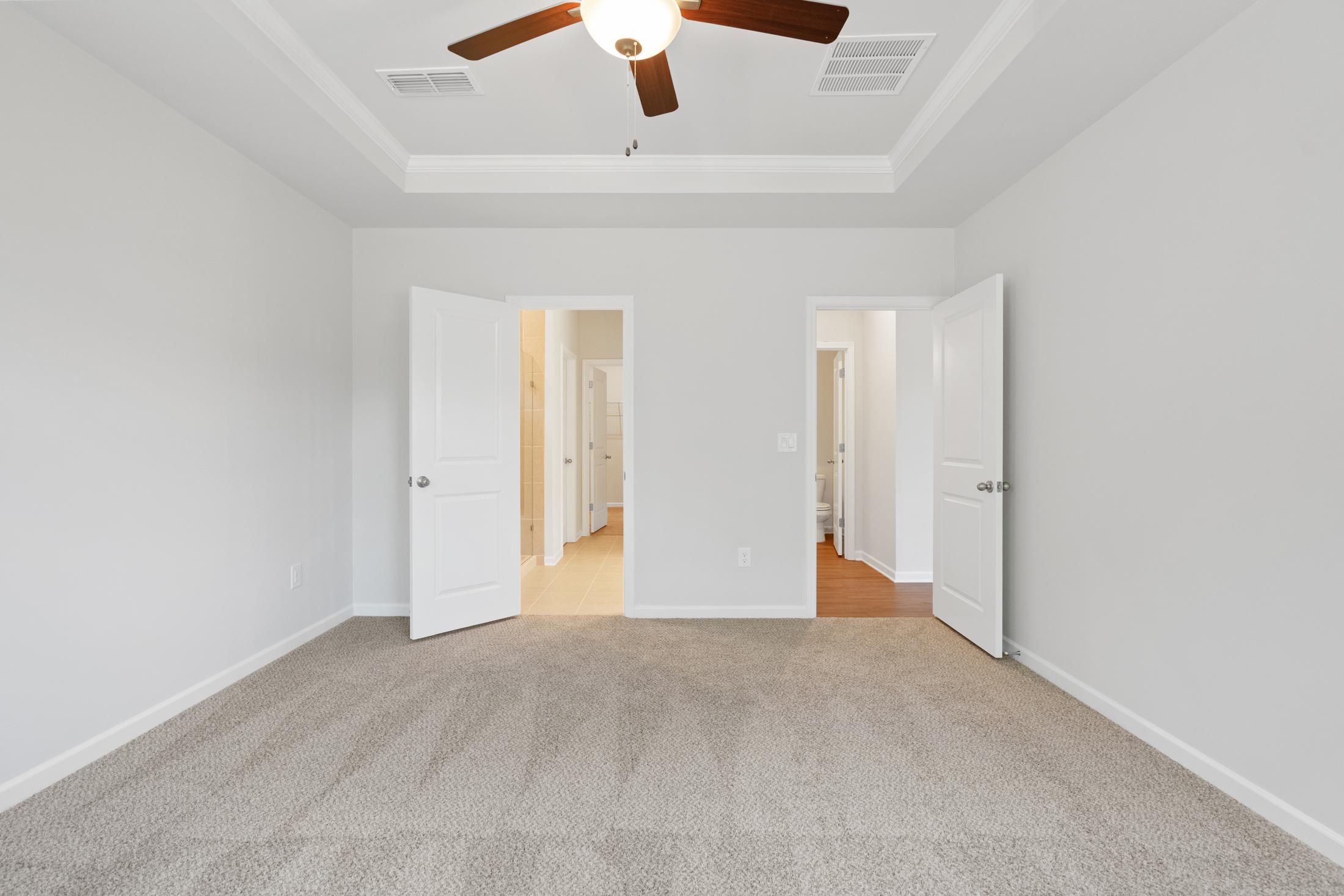 Spacious bedroom interior at Mountainbrook in Cartersville Georgia with tray ceiling fan, beige carpet, and open doors to ensuite bath and closet