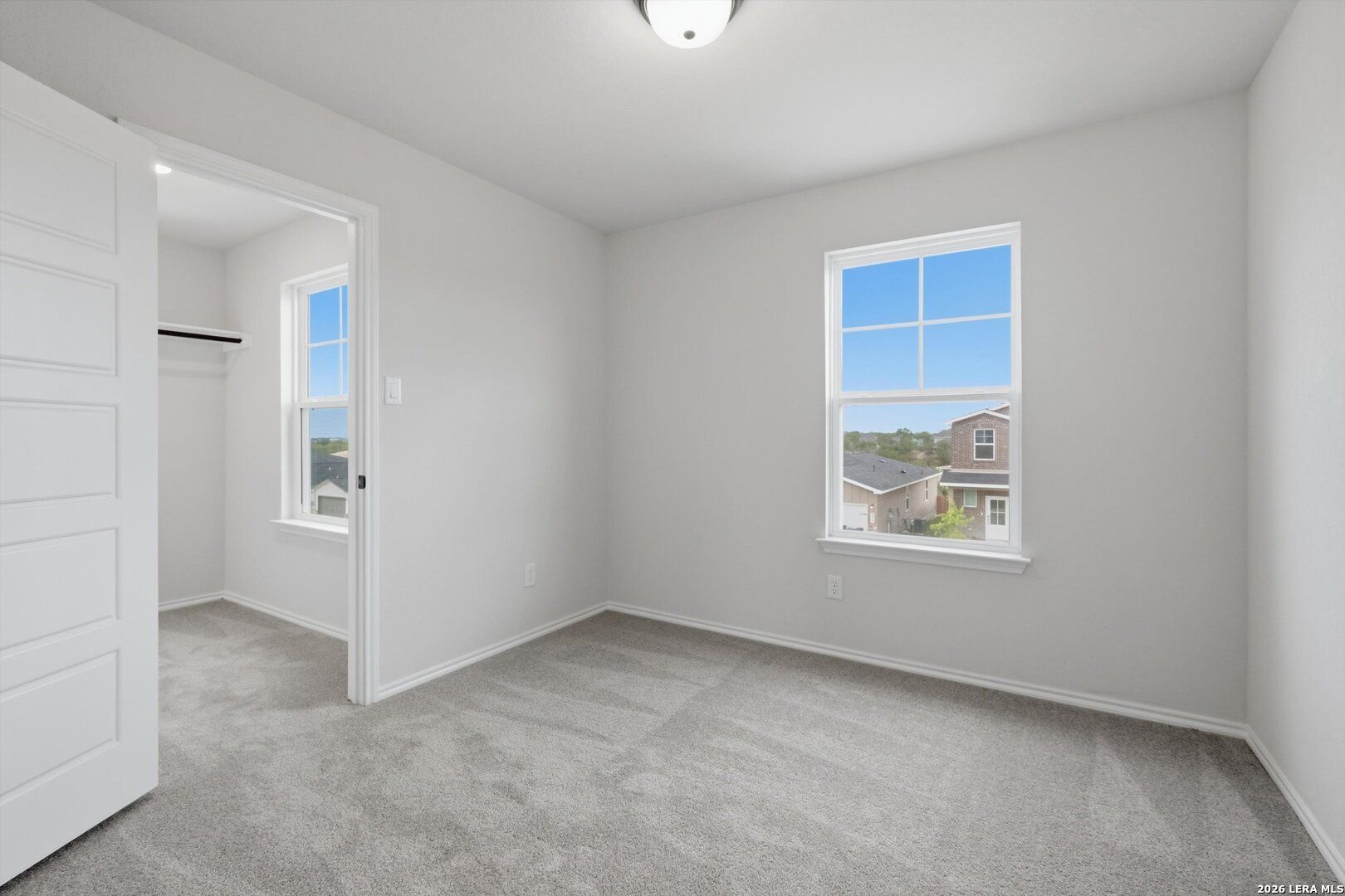 Bright bedroom with gray carpet, light gray walls, open closet door, and large window with neighborhood view in Davidson Homes The Blanco C, San Antonio