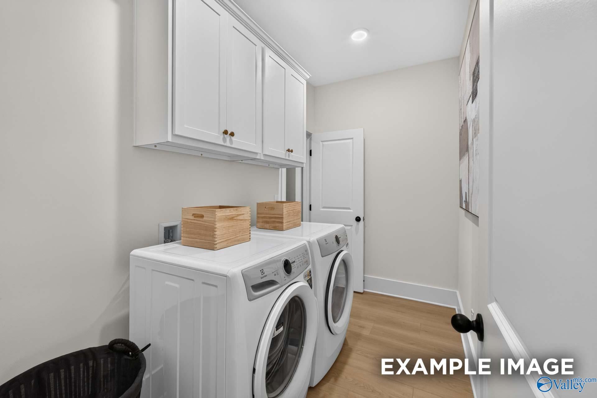 Bright laundry room with white washer and dryer, overhead cabinets, and wood-look flooring in Davidson Homes The Ashport K, Hazel Green